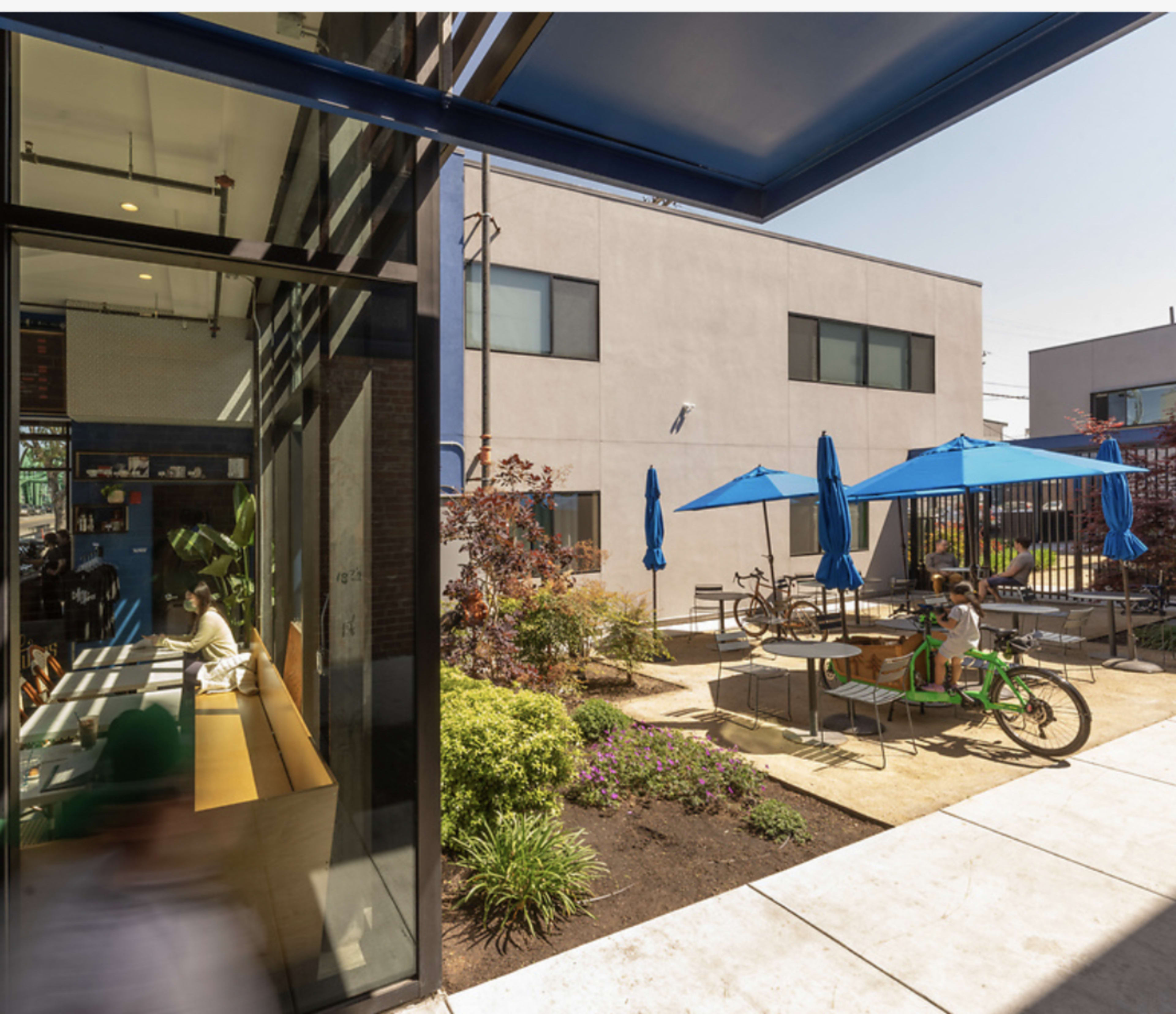 A modern outdoor seating area with blue umbrellas, bikes parked nearby, and a view of a building's glass wall reflecting a green interior space.