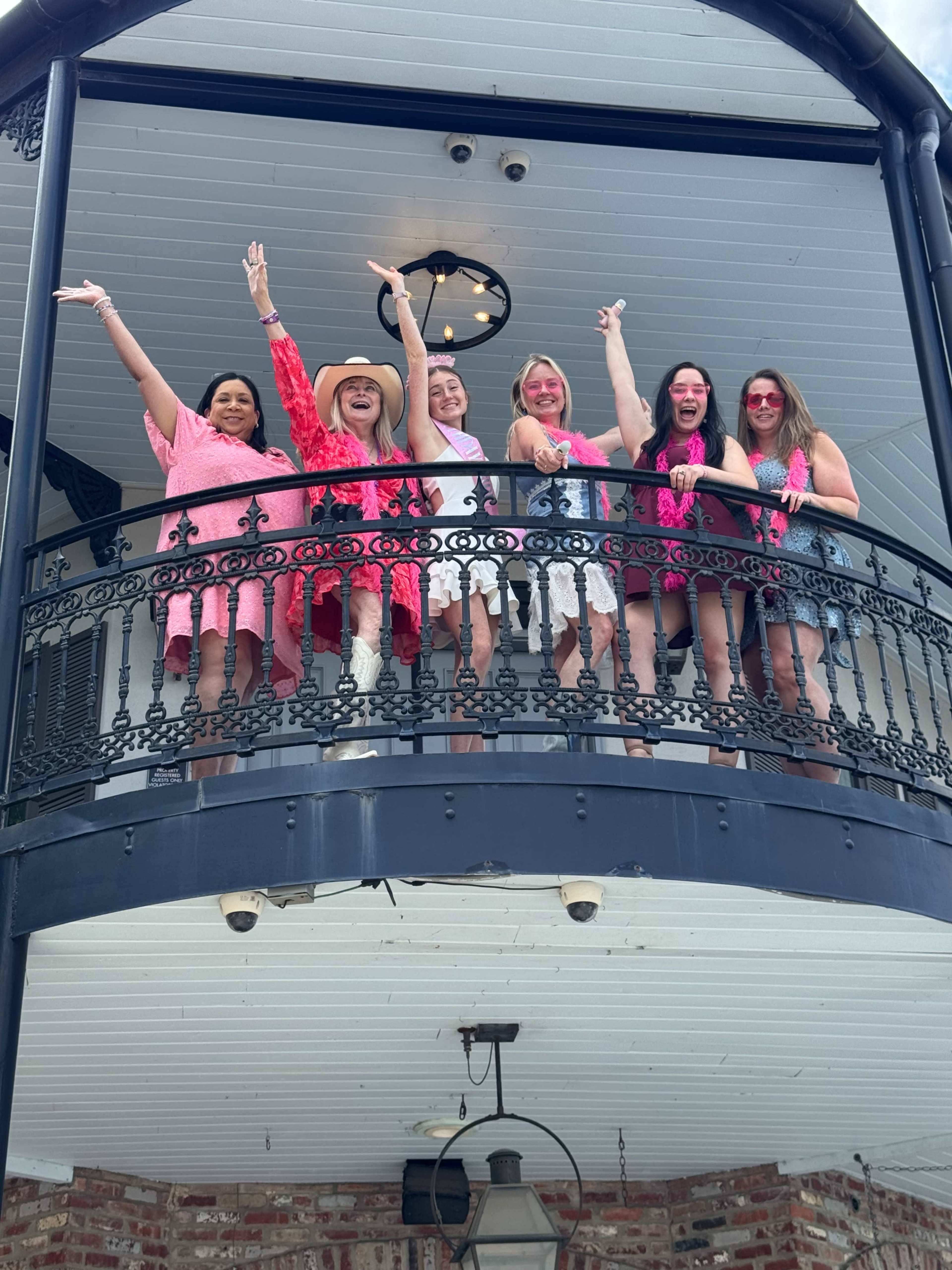 A group of six women stands on a balcony, smiling and posing with arms raised, dressed in bright clothing.