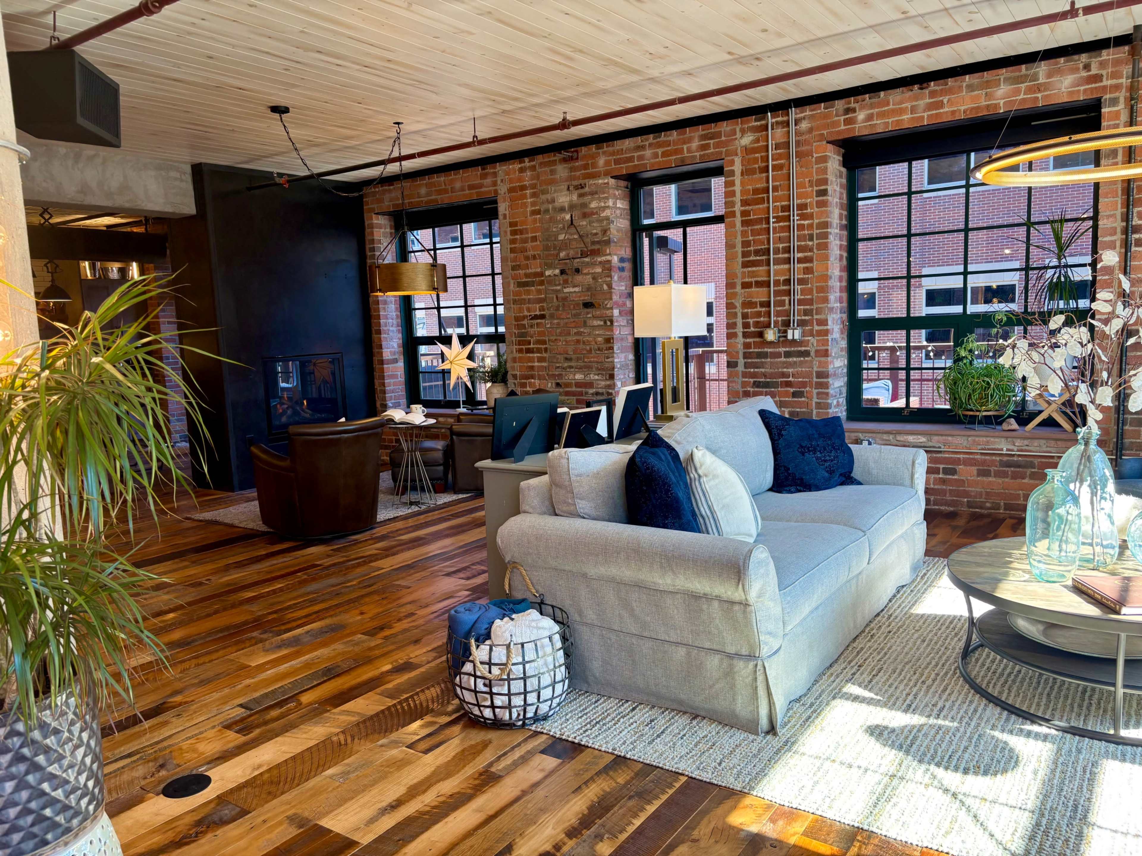 The image shows a stylish living area with a gray sofa, wooden floors, brick walls, and large windows, featuring decorative plants and modern furnishings.