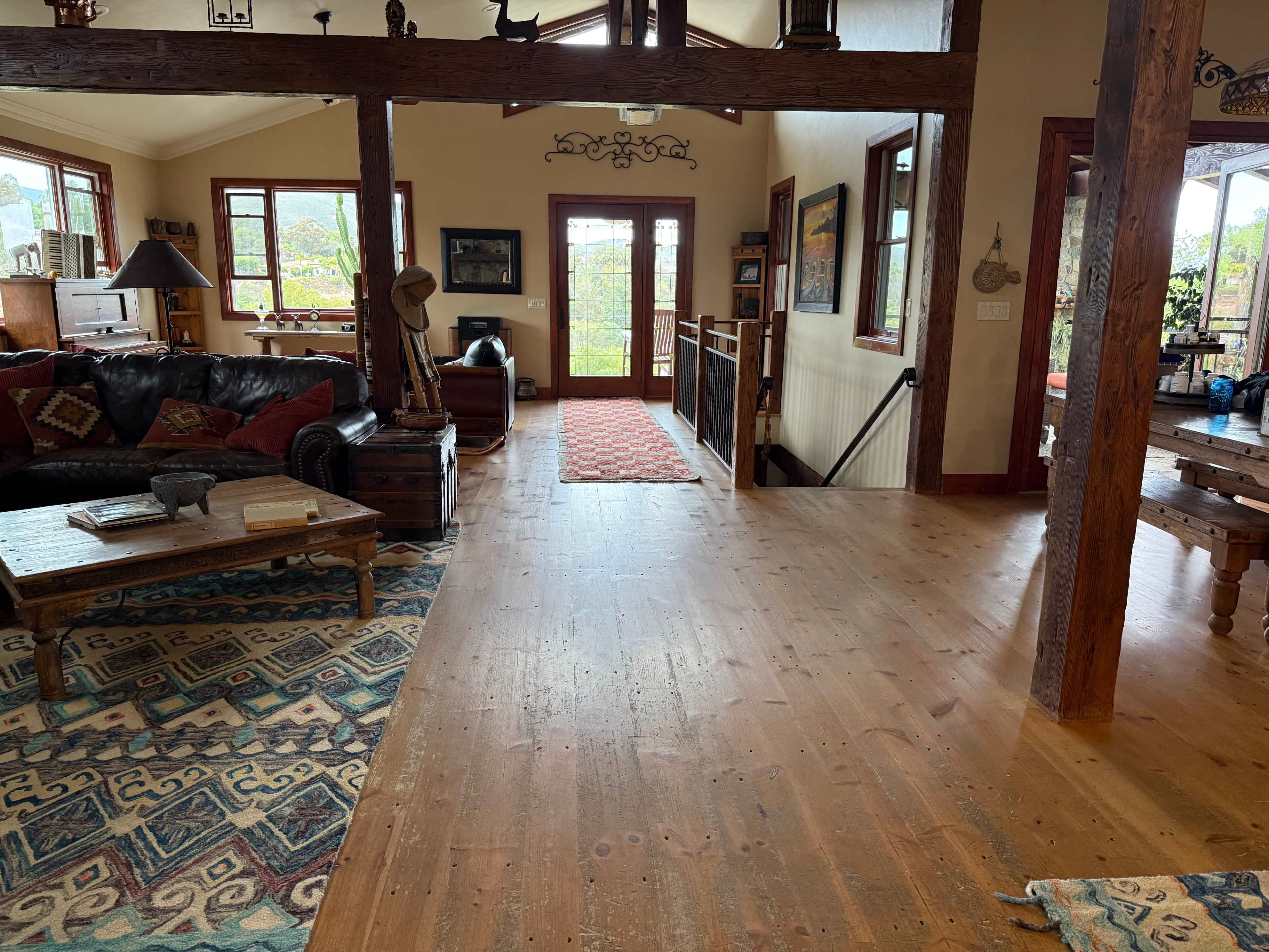 The interior of a cozy home features wooden flooring, a leather couch, and a decoratively patterned rug, with large windows allowing natural light to enter.