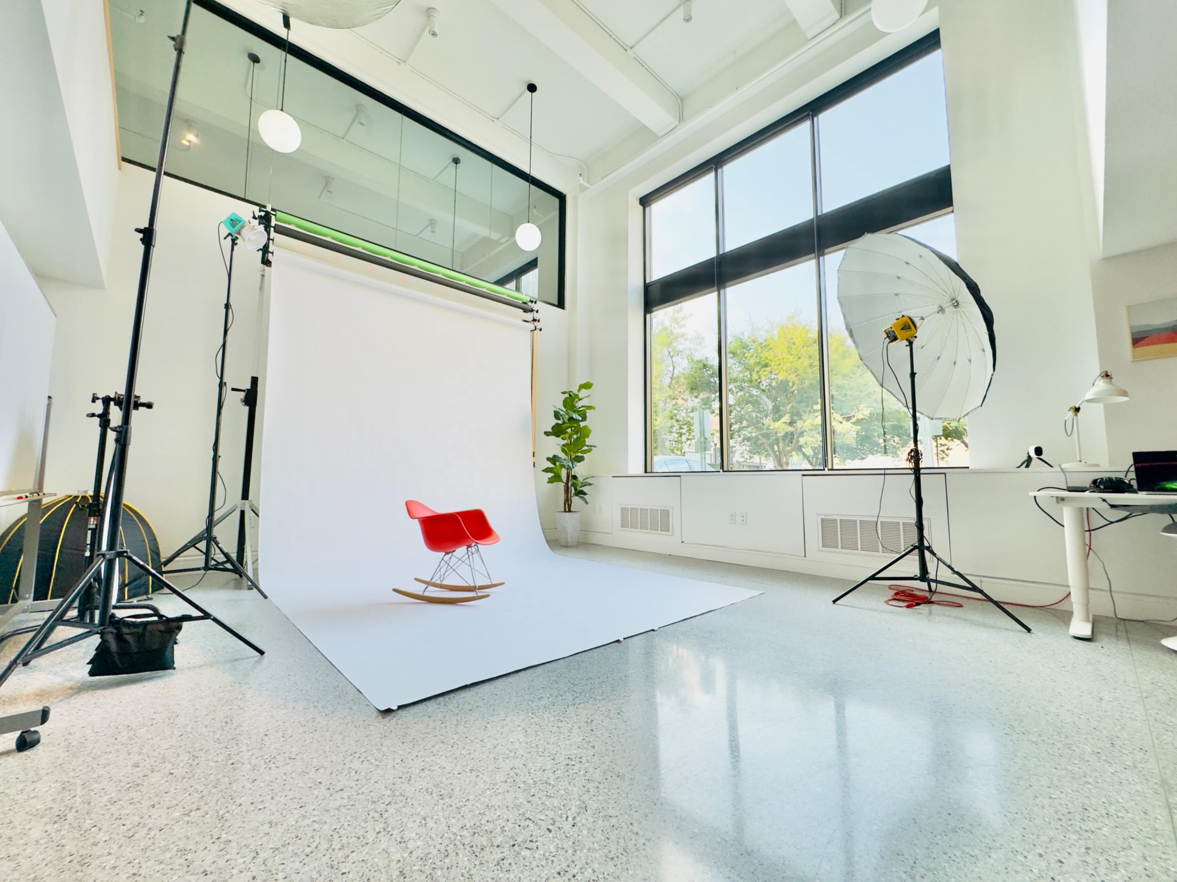 The image shows a bright photo studio featuring a red rocking chair against a white backdrop, with studio lights and a green plant nearby.