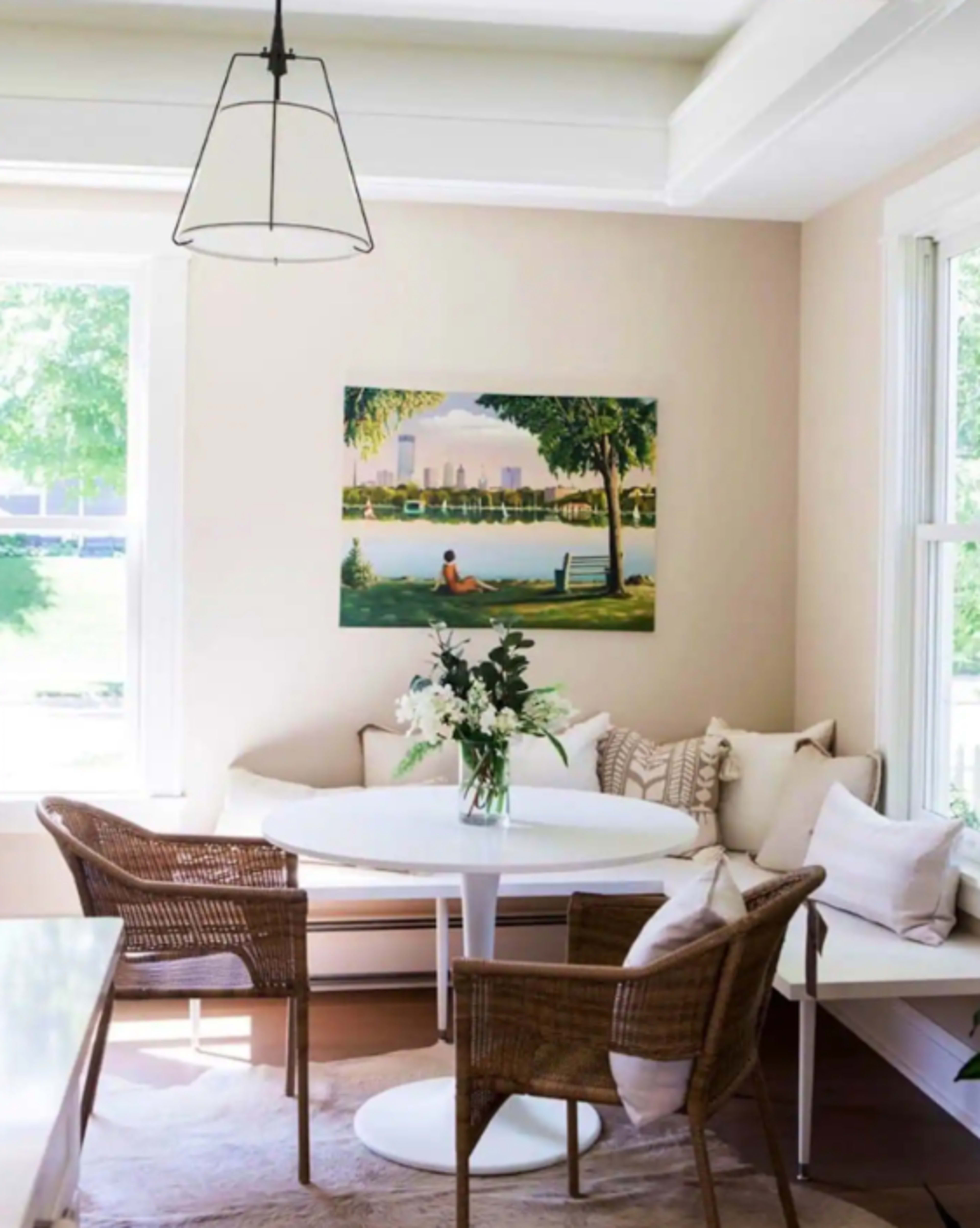 A dining area features a round white table surrounded by wicker chairs and adorned with a vase of flowers, with a large painting of a park scene on the wall.