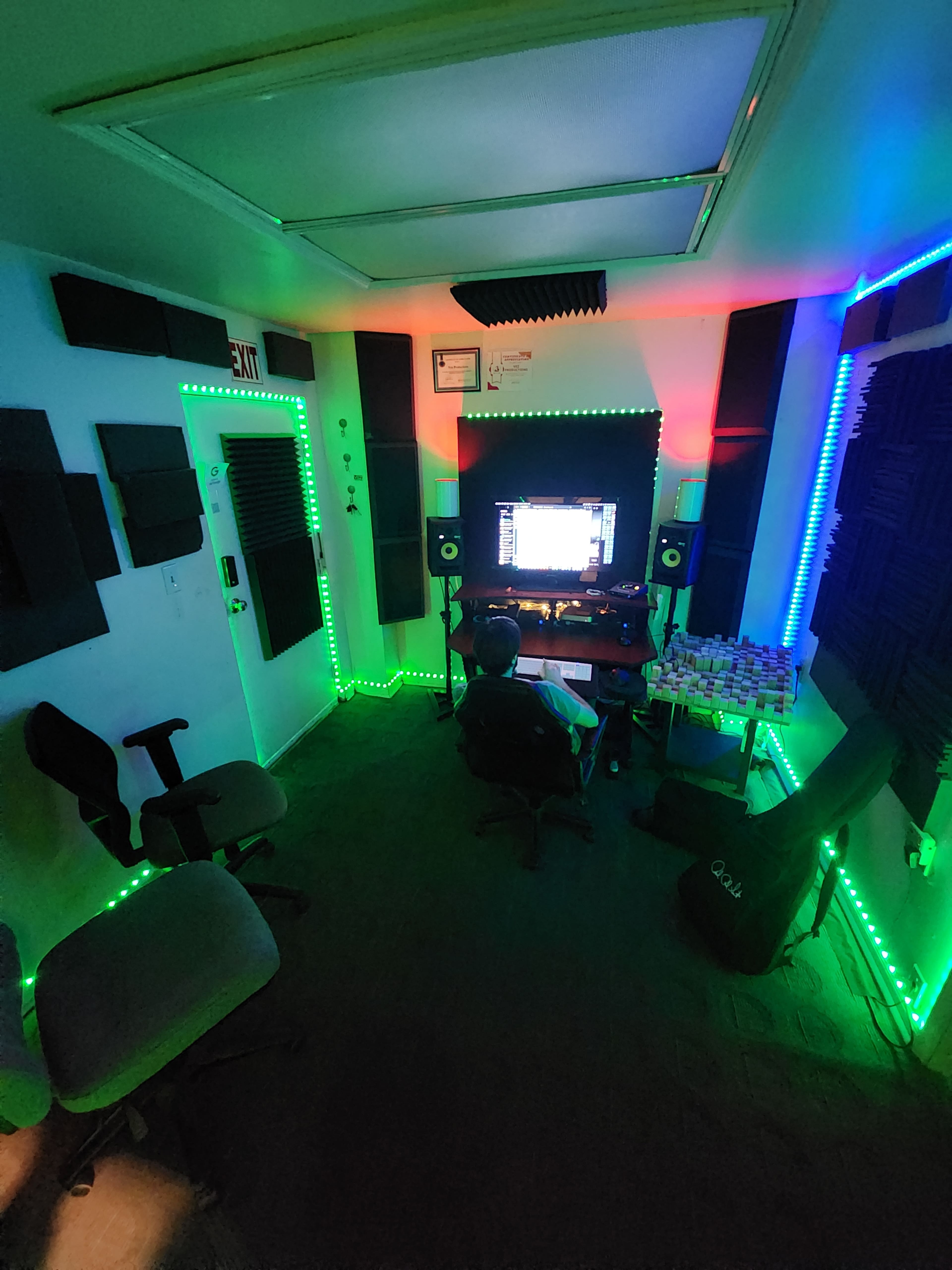 A person sits in a chair in a music studio, surrounded by soundproofing panels and illuminated by colorful LED lights while working on a computer.