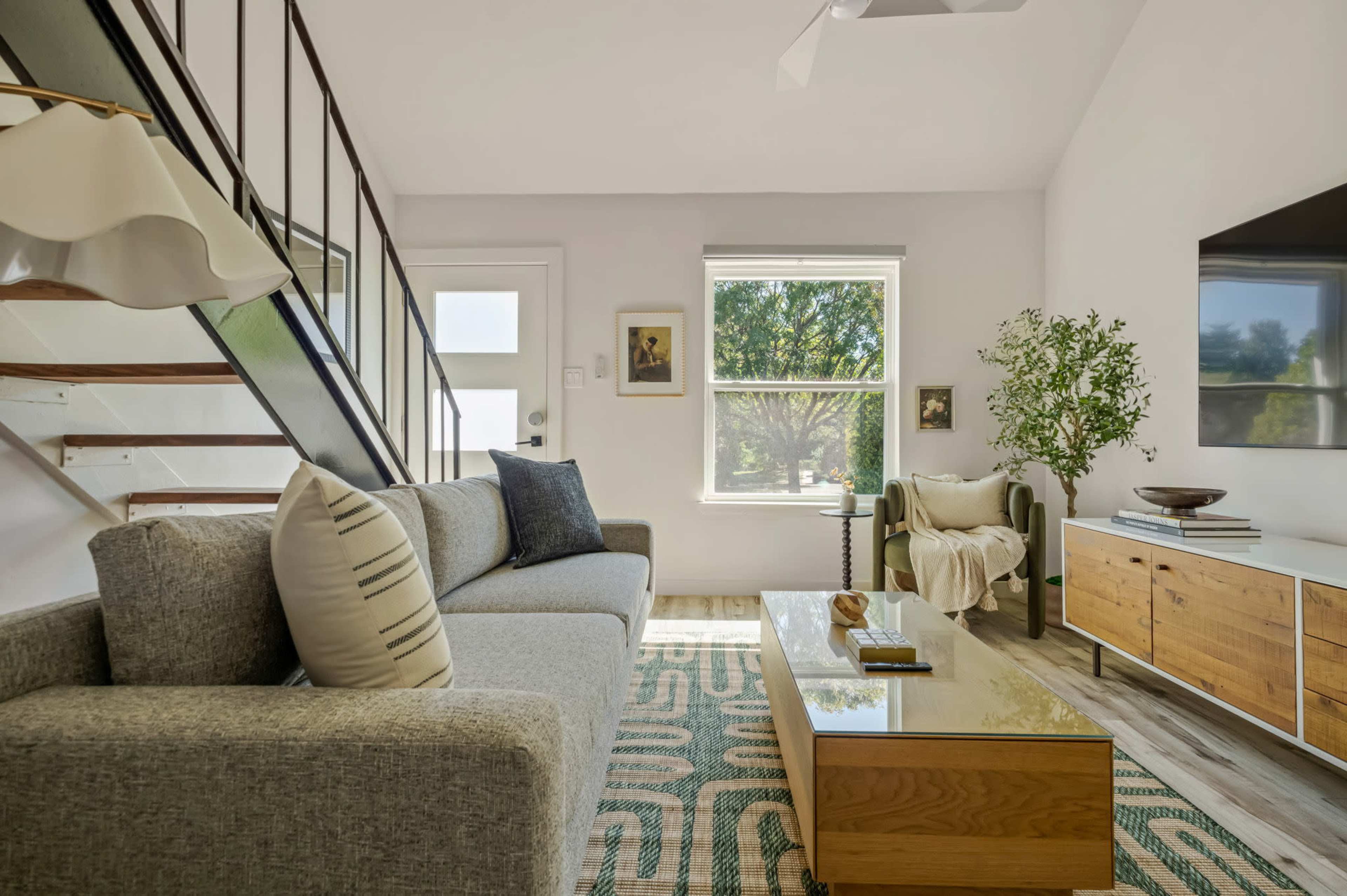 A modern living room features a gray sofa, a wooden coffee table, and a green accent chair, with large windows allowing natural light to fill the space.