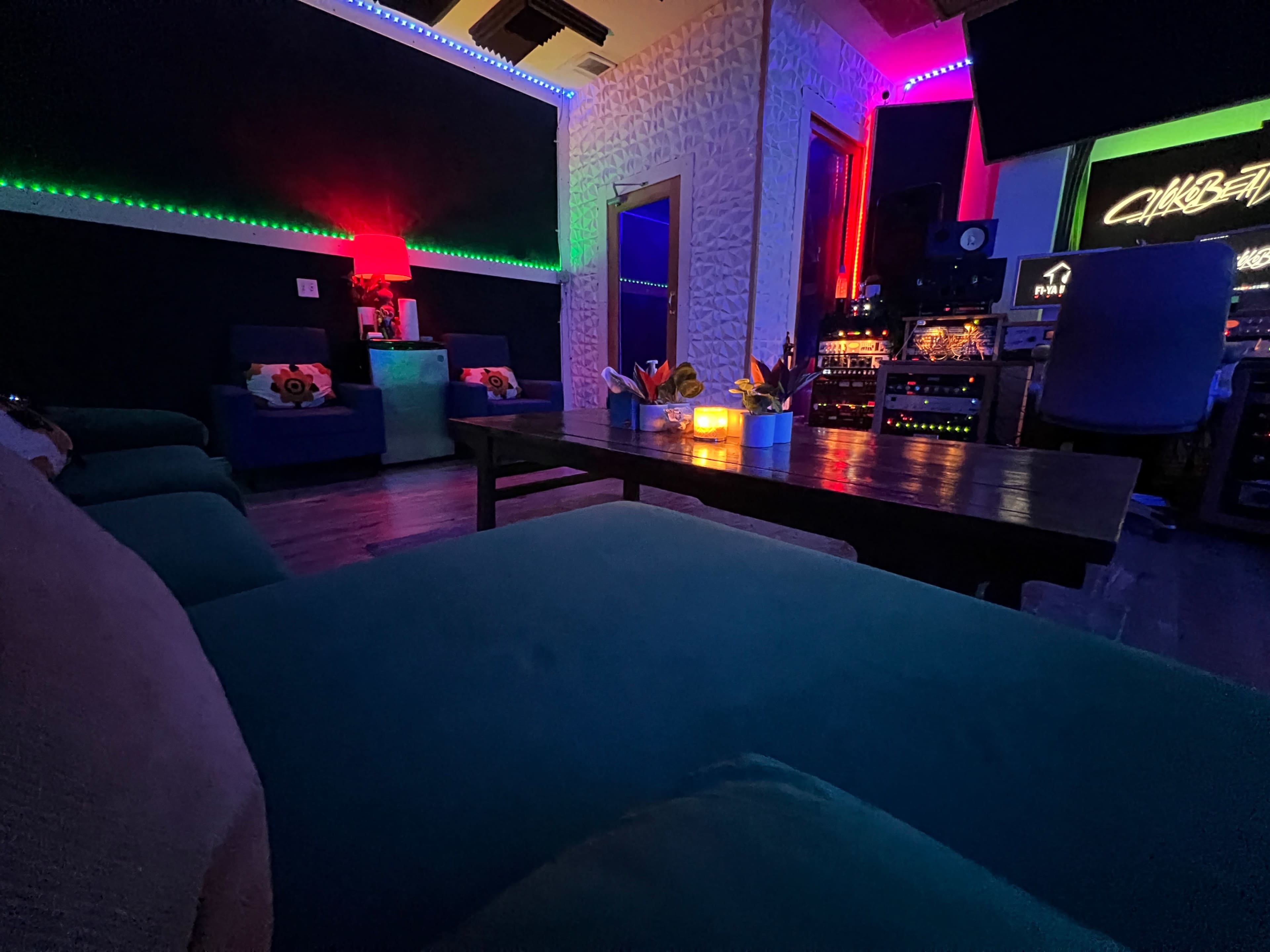 The image shows a dimly lit recording studio with colorful LED lights, a wooden table at the center, and sound equipment visible in the background.