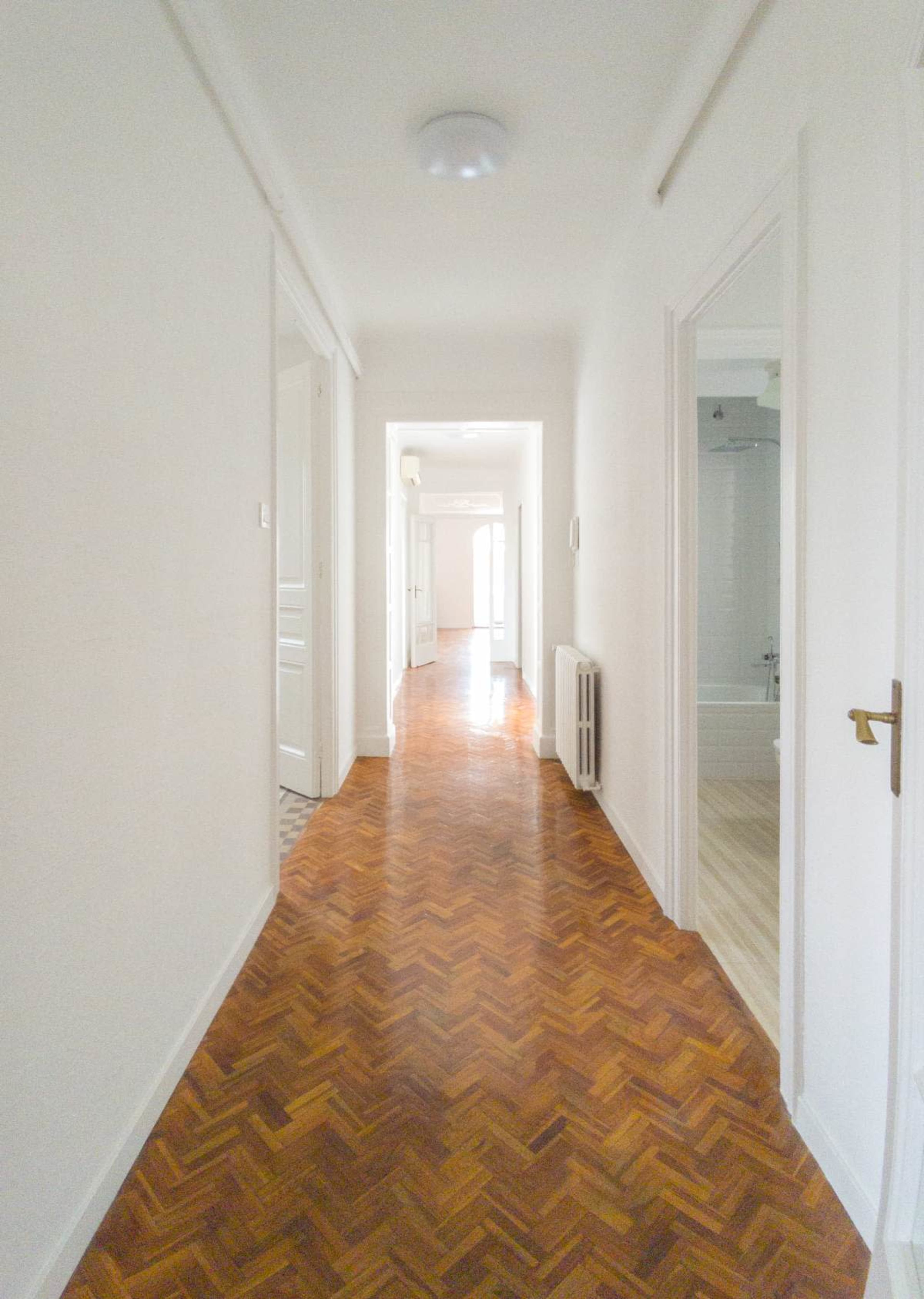 A long, narrow hallway with wooden herringbone flooring and white walls leads to multiple doorways.
