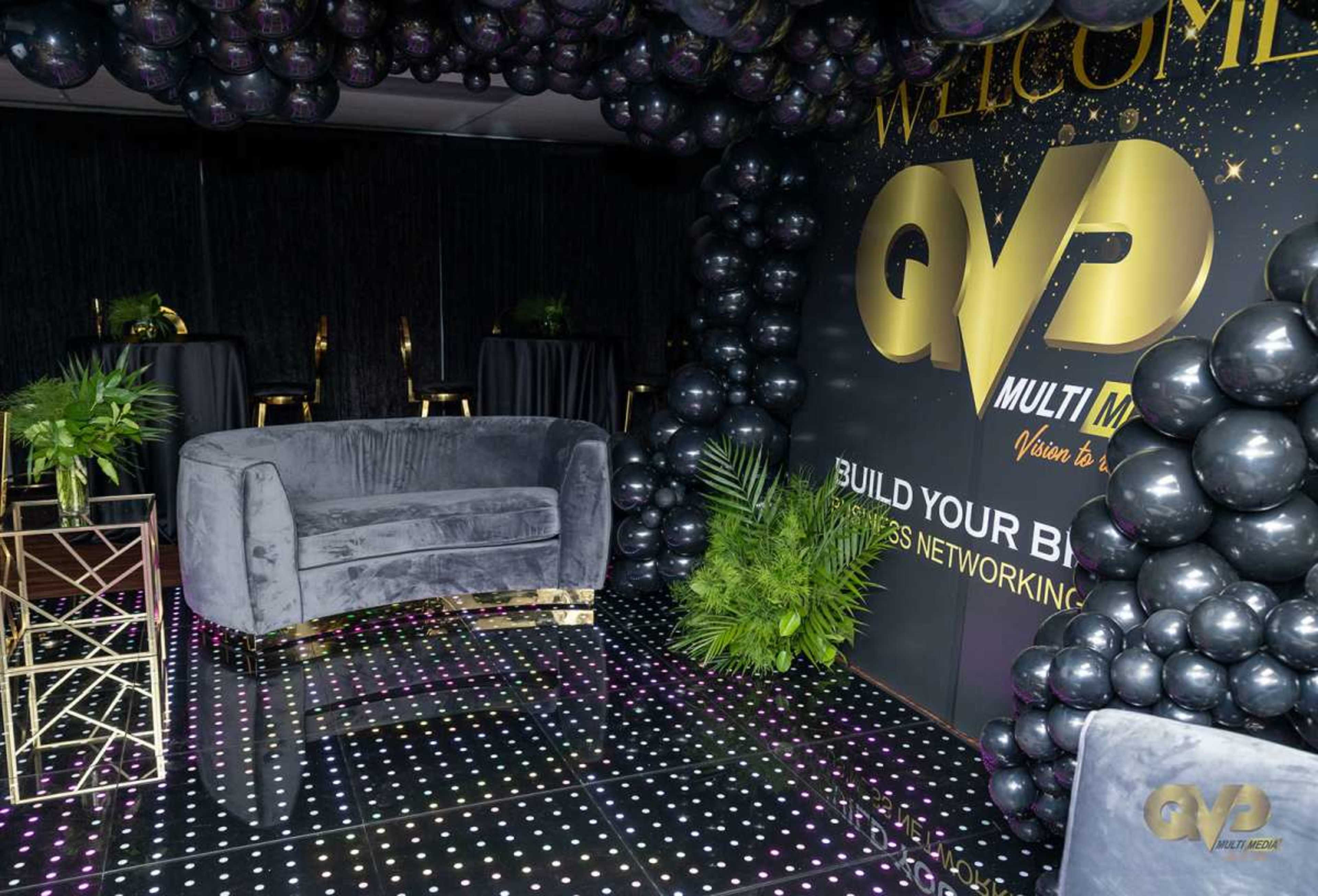 The image shows a stylish event space decorated with black and gold balloons, plants, a velvet sofa, and high tables.