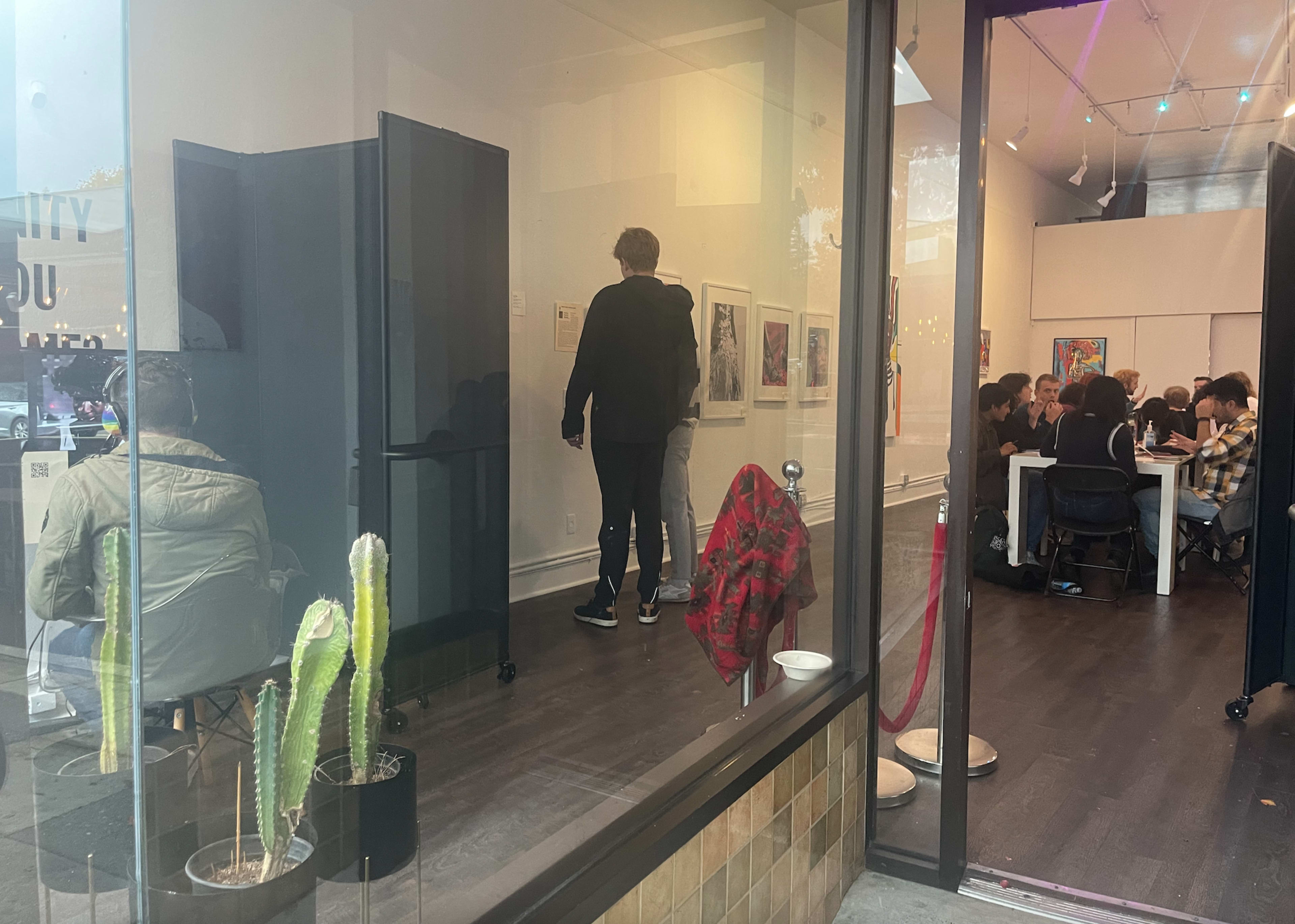 A gallery setting with people viewing artwork on the walls while a group is seated at a table in the background.