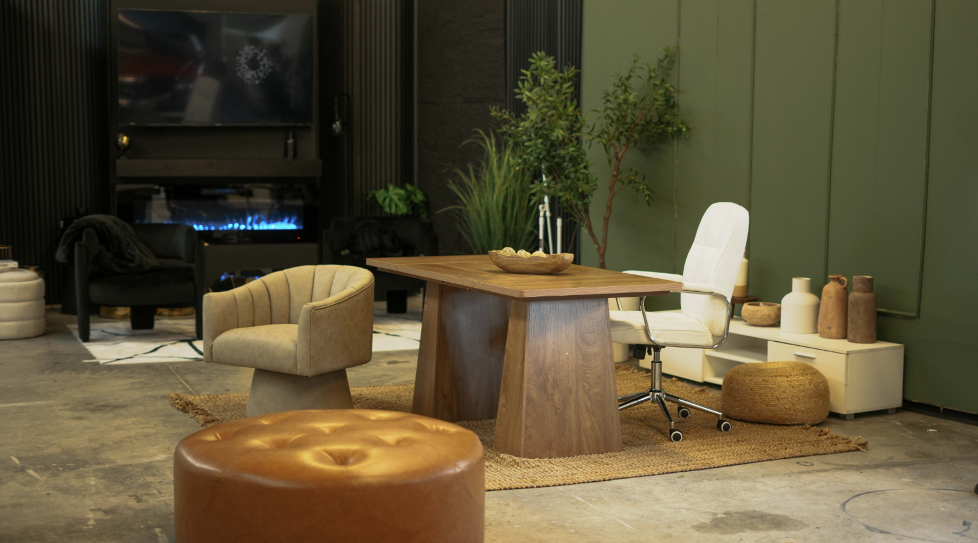 The image features a modern workspace setup with a wooden table, an office chair, a decorative plant, and various seating options arranged on a textured rug.