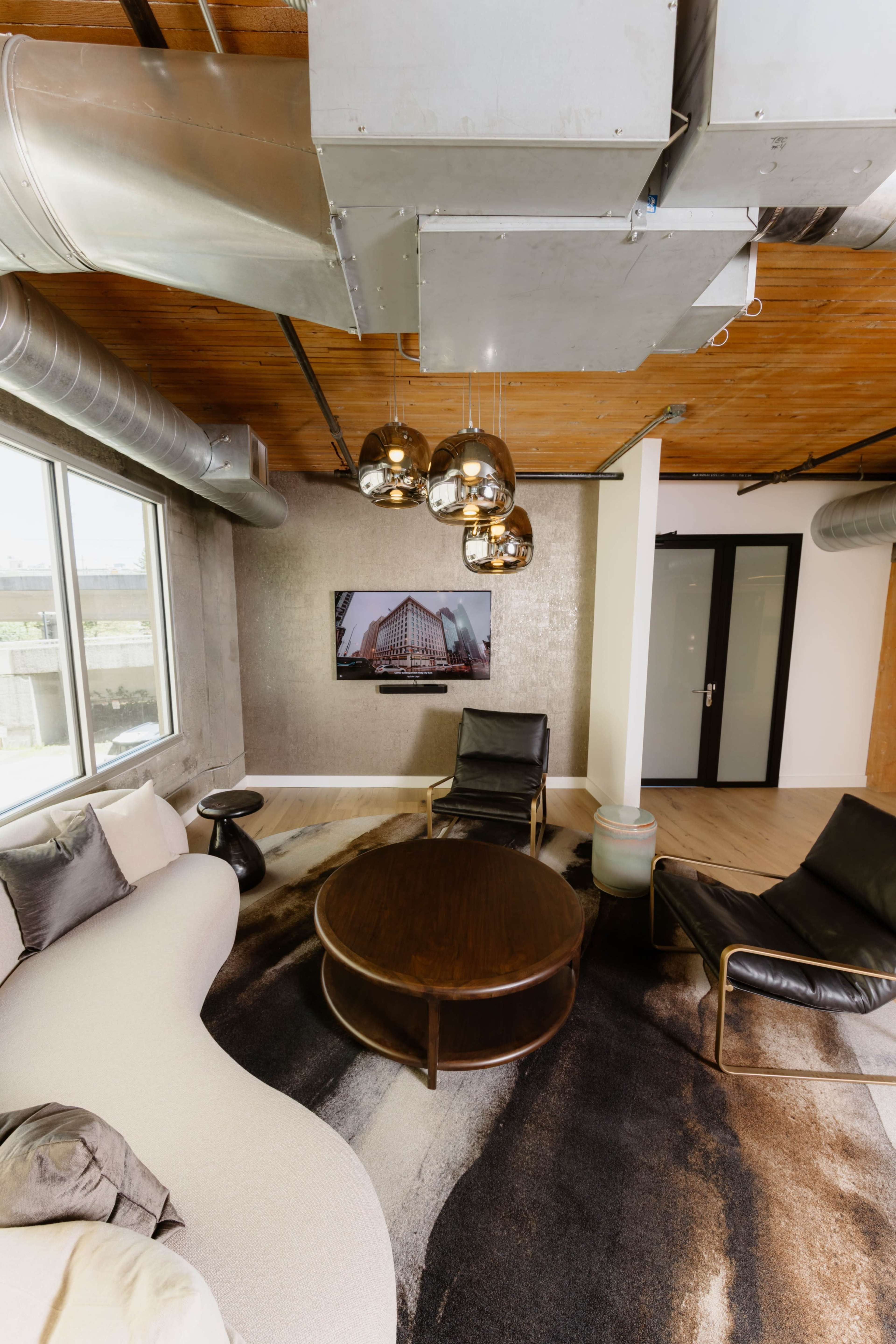 The image shows a modern living space featuring a curved white sofa, two black chairs, a round wooden coffee table, and a television mounted on the wall, with industrial-style ductwork exposed overhead.