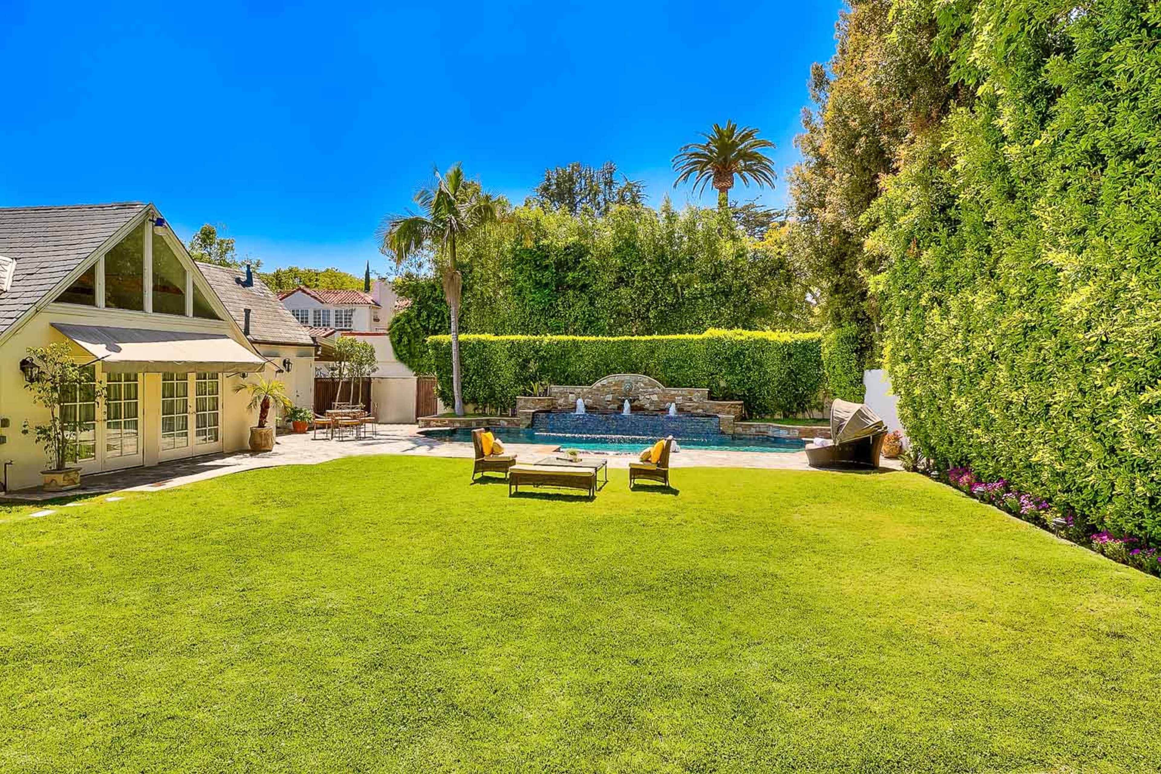 A landscaped backyard with a swimming pool, a stone fountain, and lounge chairs set on a manicured lawn surrounded by tall hedges and palm trees.