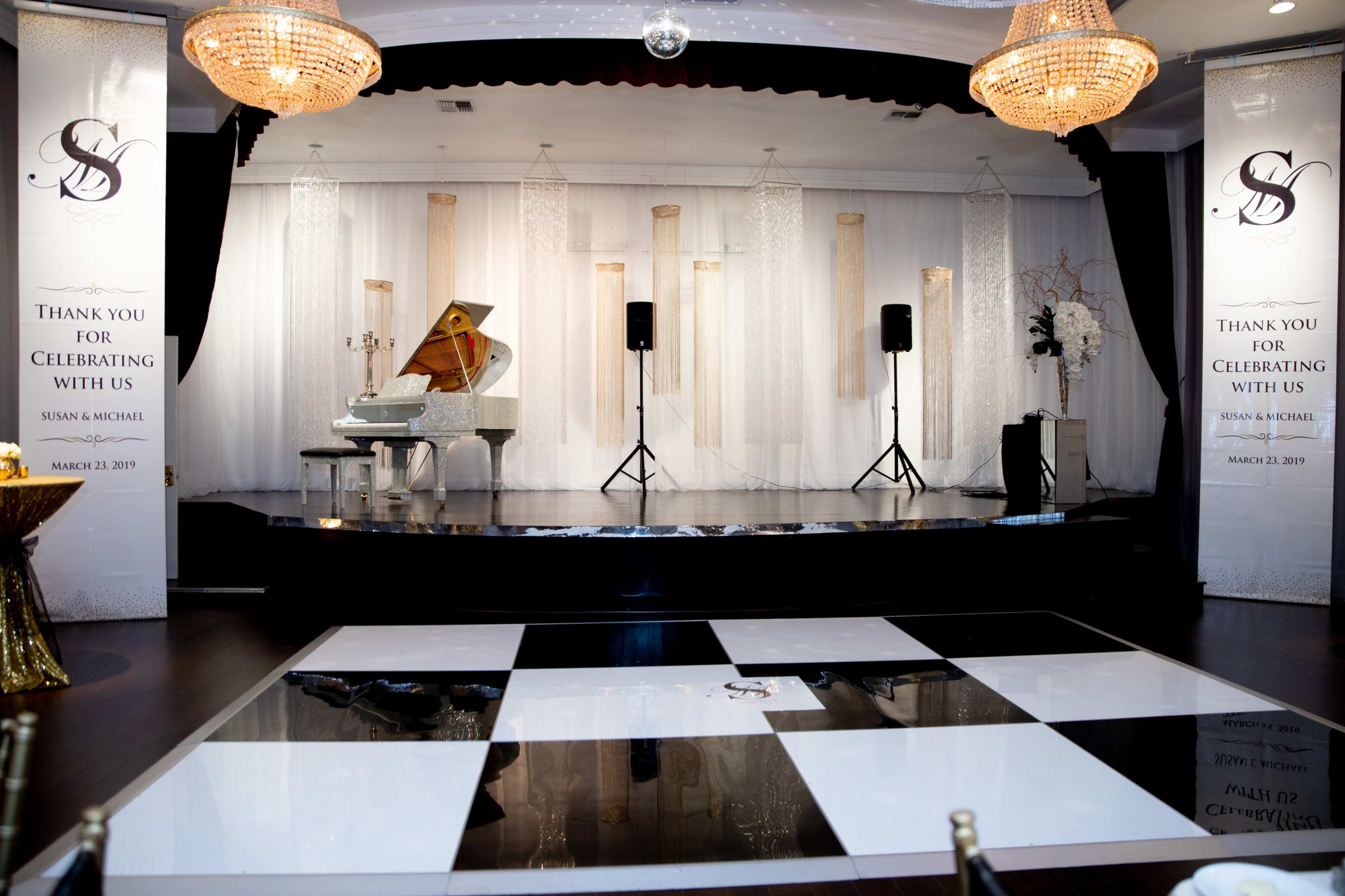 A elegantly decorated event space featuring a grand piano on a stage, black and white dance floor tiles, and large banners thanking guests for their attendance.