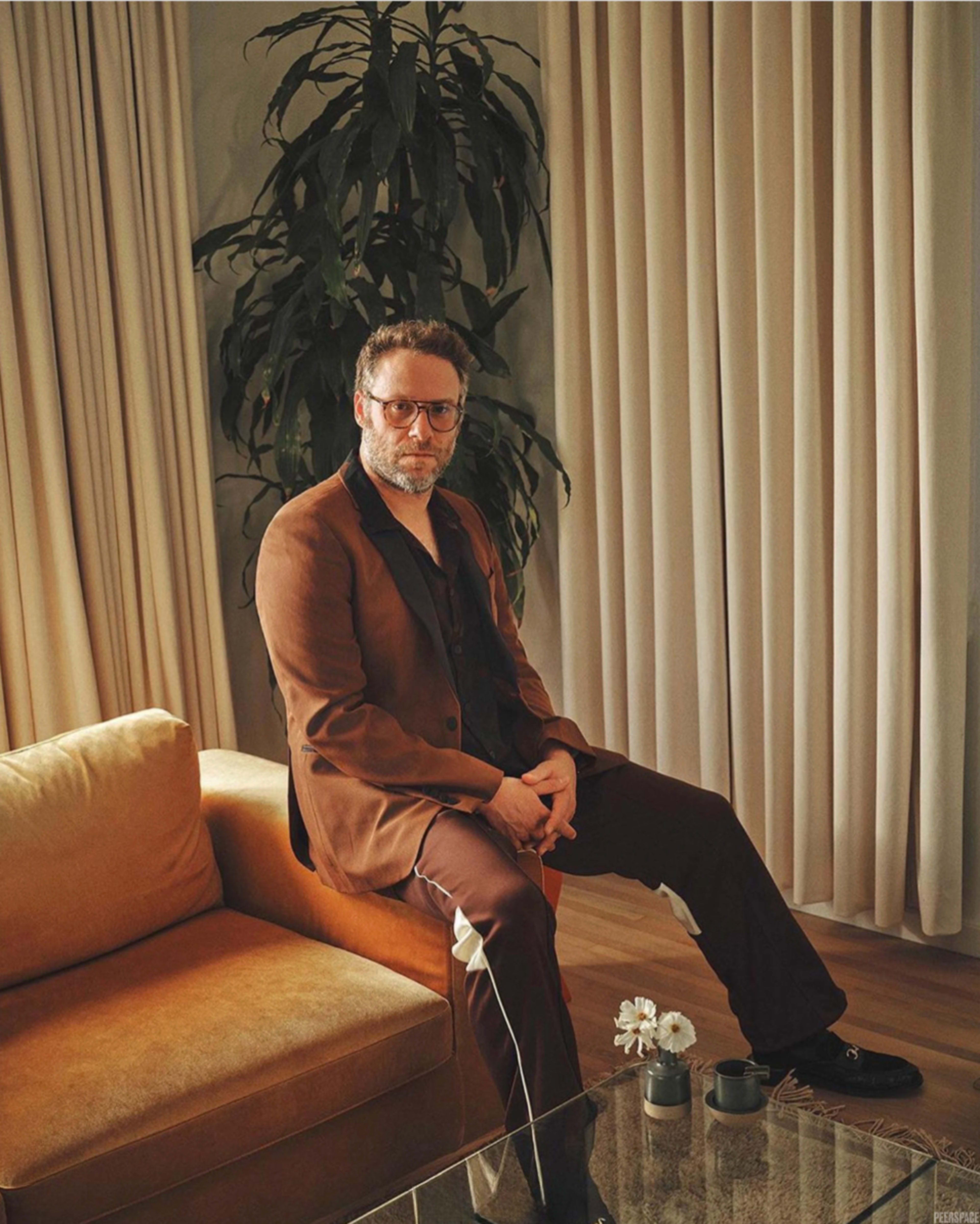 A man sits on a brown couch in a well-lit room with large curtains and a potted plant nearby.