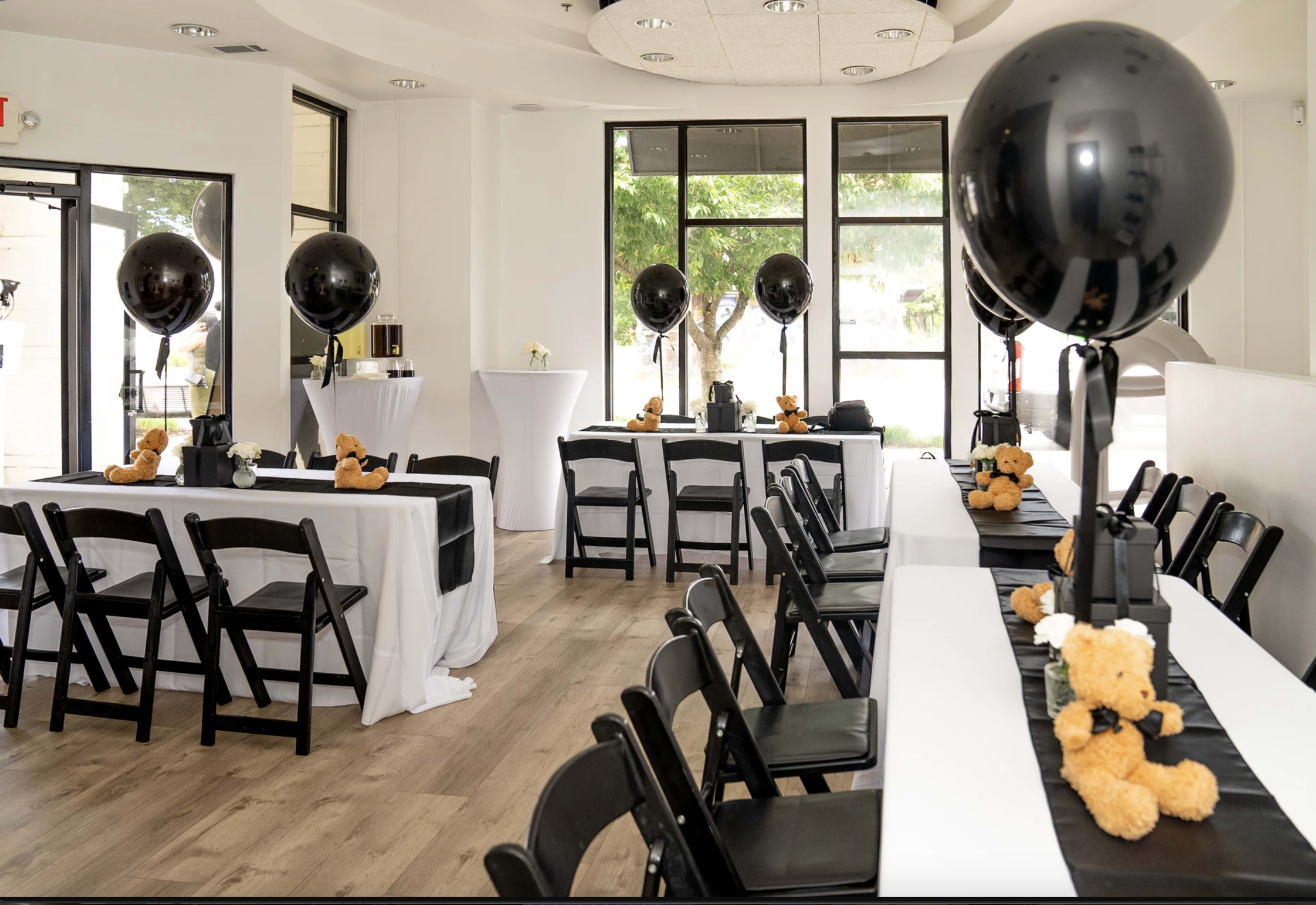 A room is set up for a gathering with black balloons and teddy bears placed on white tablecloths.
