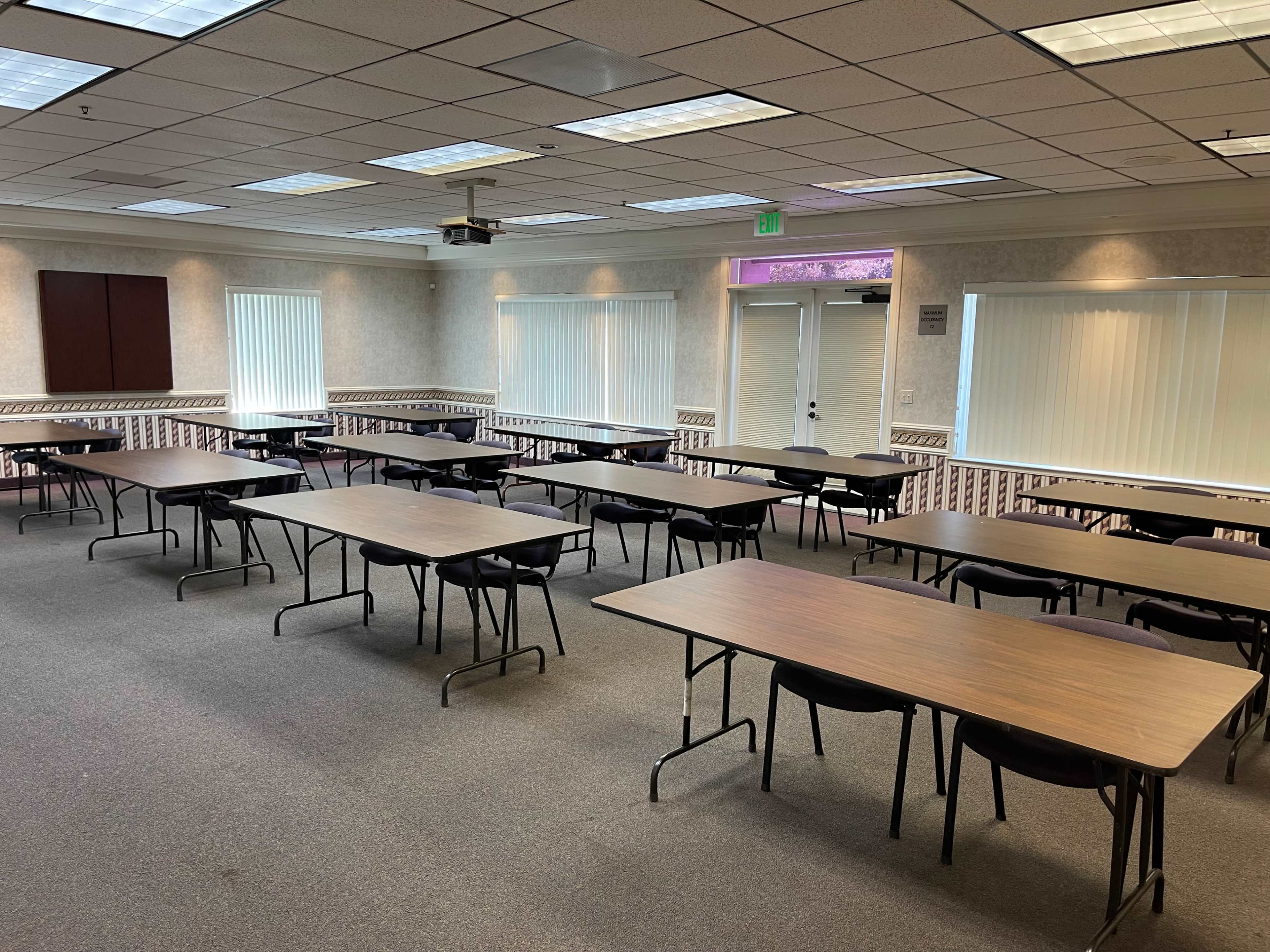 A meeting room arranged with several rows of tables and chairs, all facing a projector screen at the front.