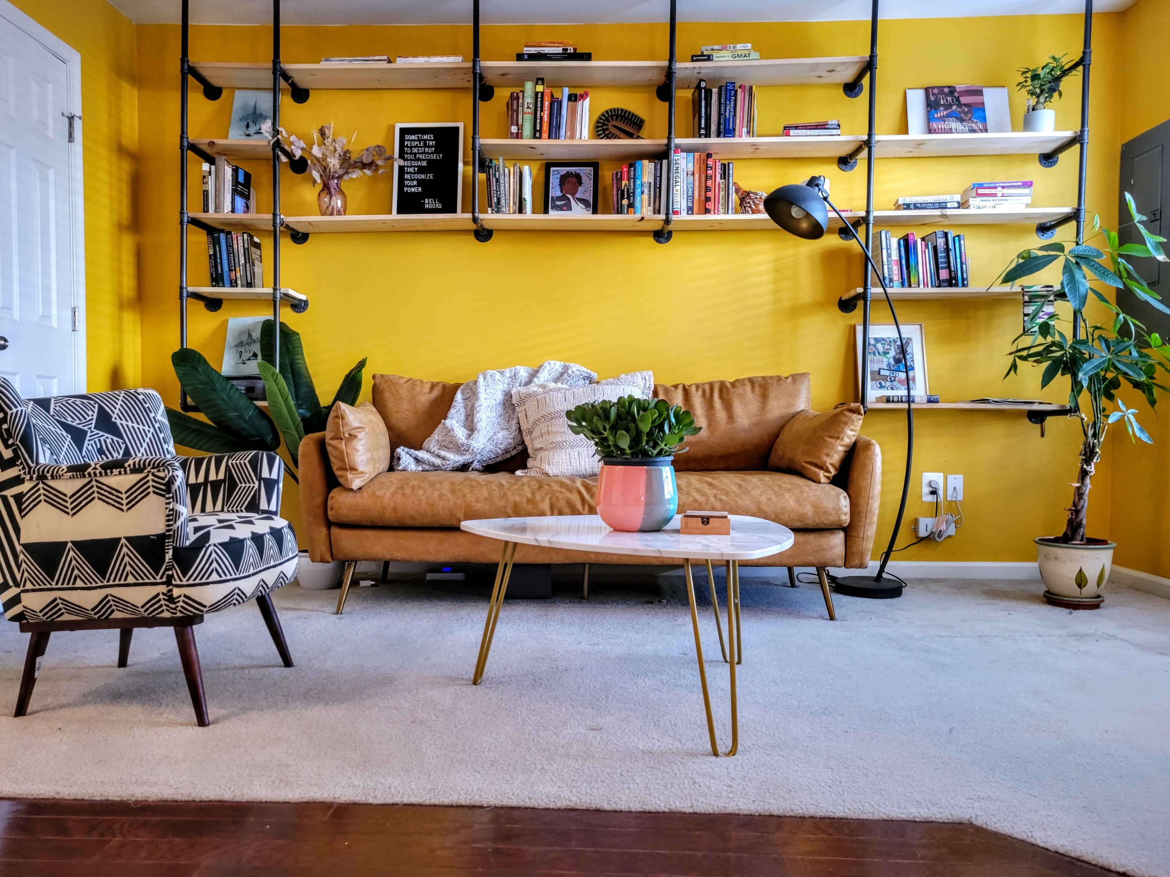 The living room features a yellow wall with bookshelves, a brown leather couch, a round coffee table, and various plants.