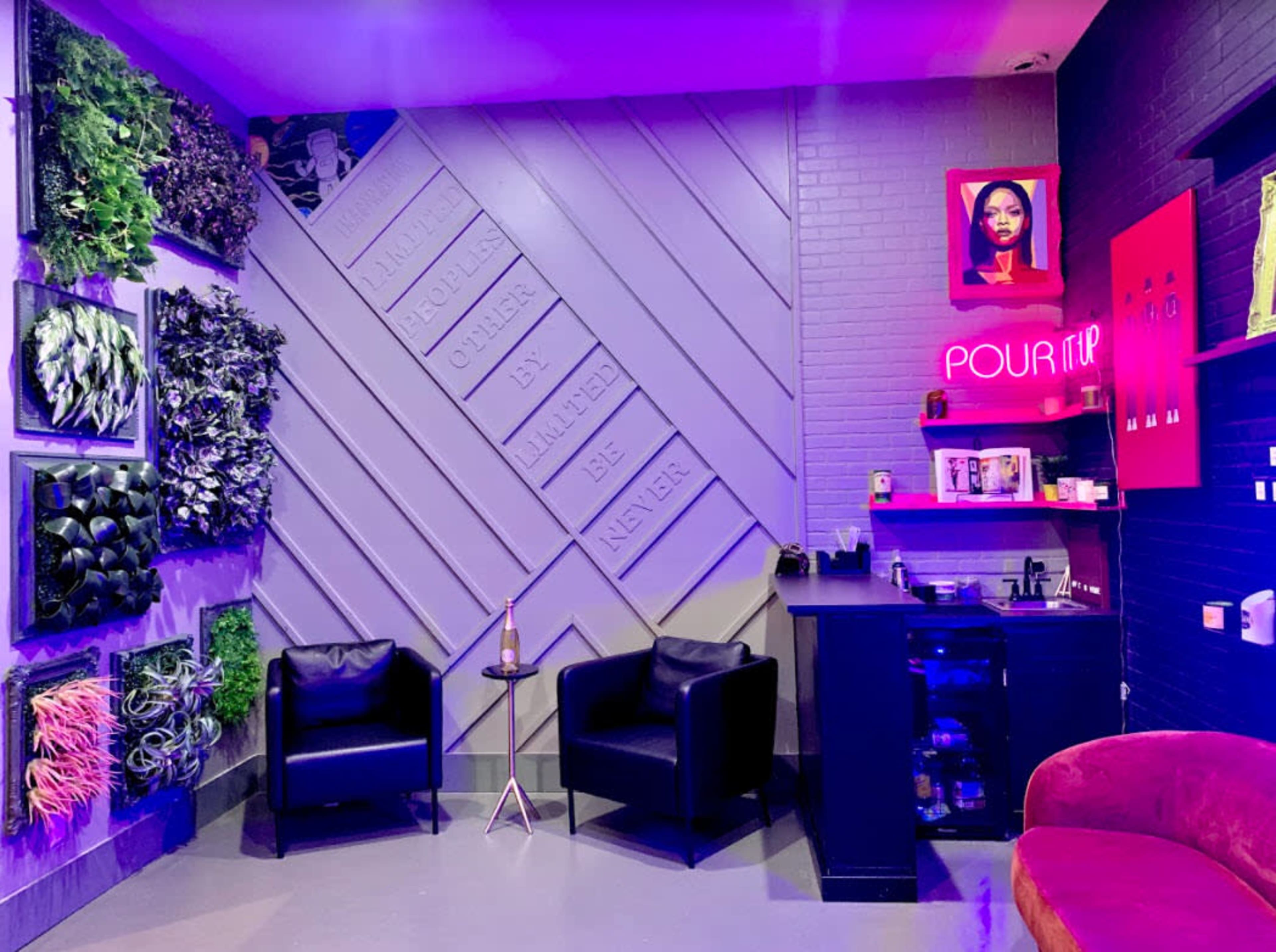 A modern interior of a lounge area with black chairs, a neon sign, and a wall adorned with framed artworks and greenery.