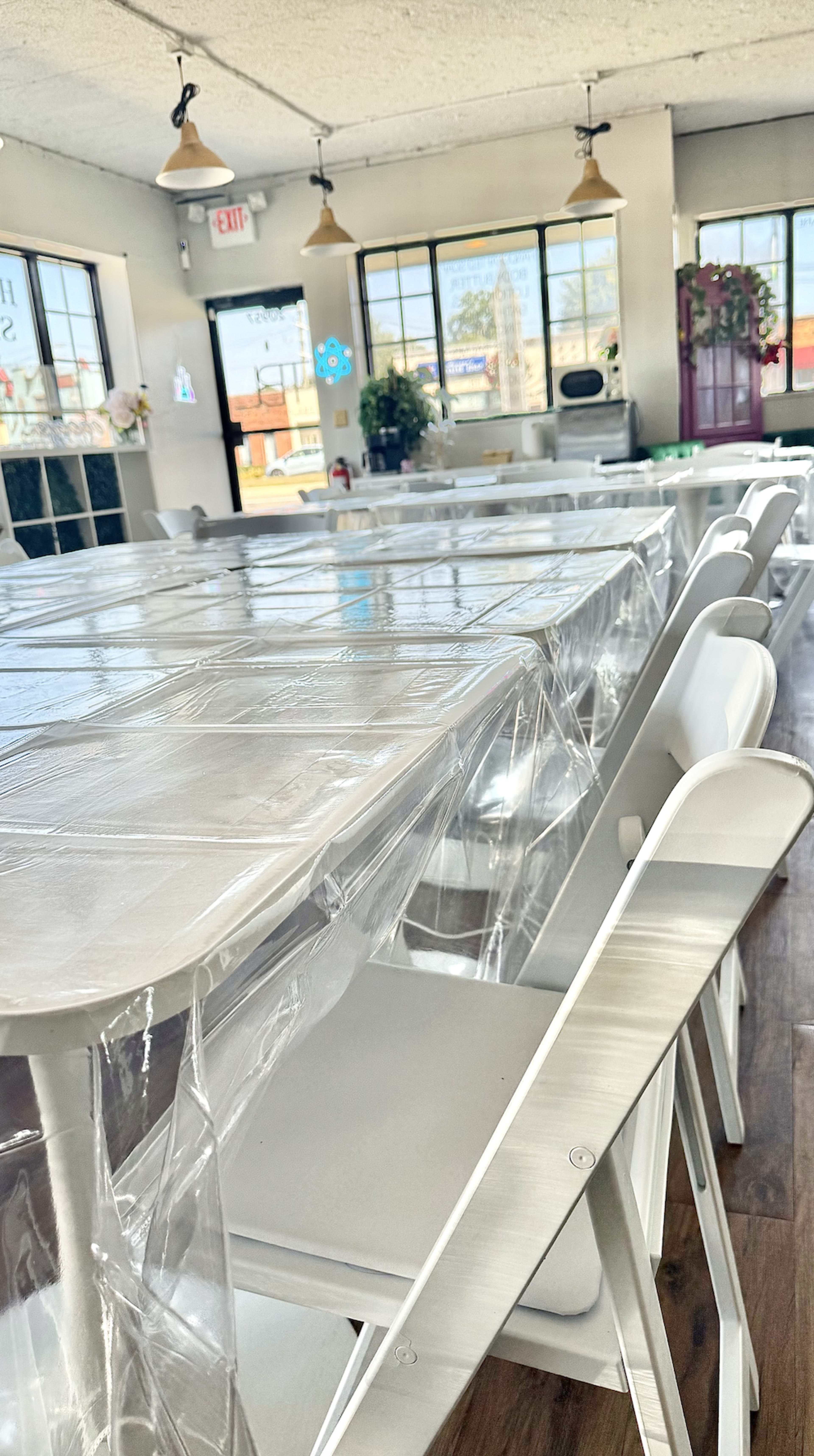 A spacious dining area features white tables and chairs, with plastic coverings on the tables, and large windows allowing sunlight to enter.