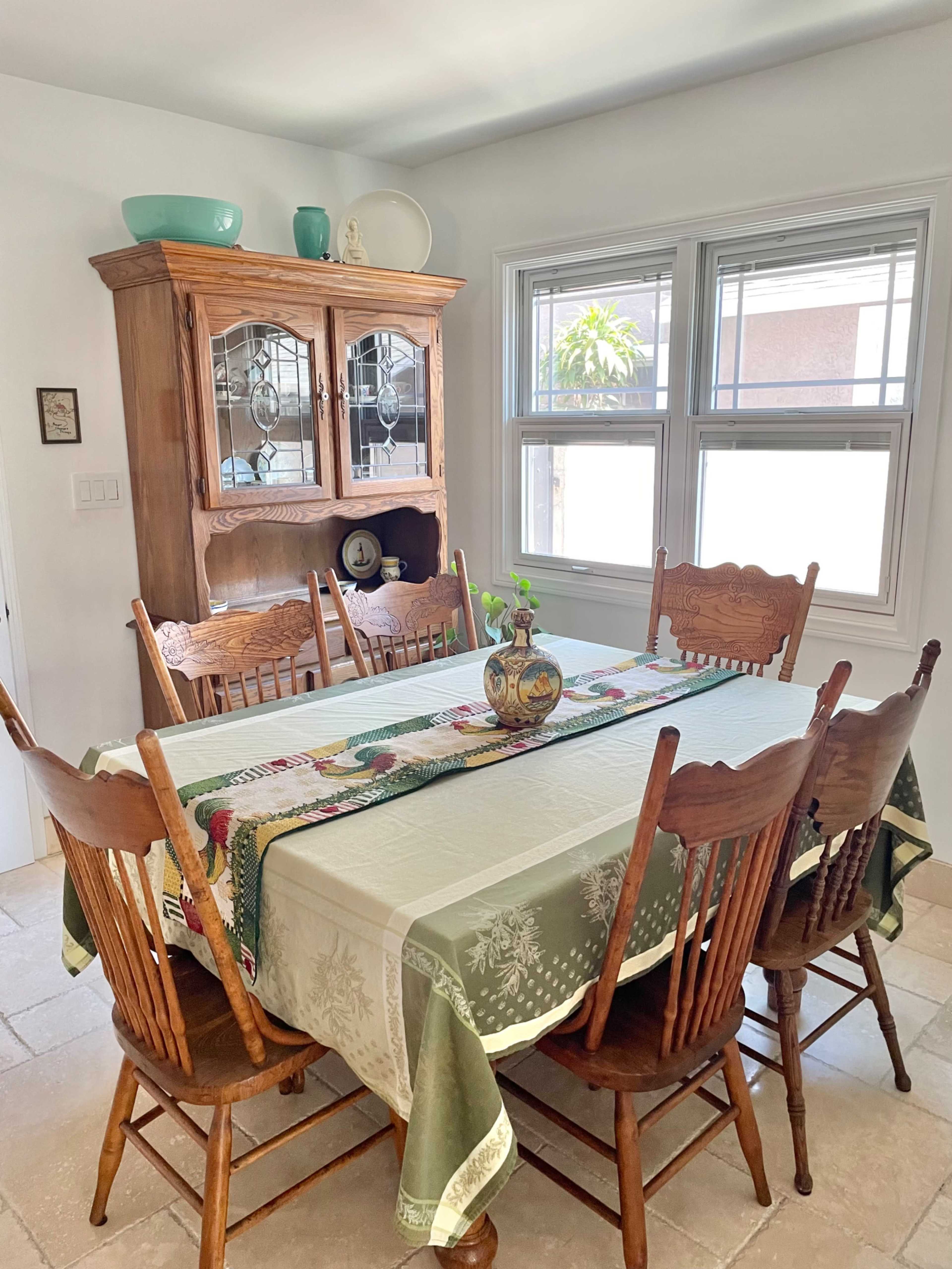 A dining room features a wooden table surrounded by eight chairs, with a hutch displaying dishes in the background.