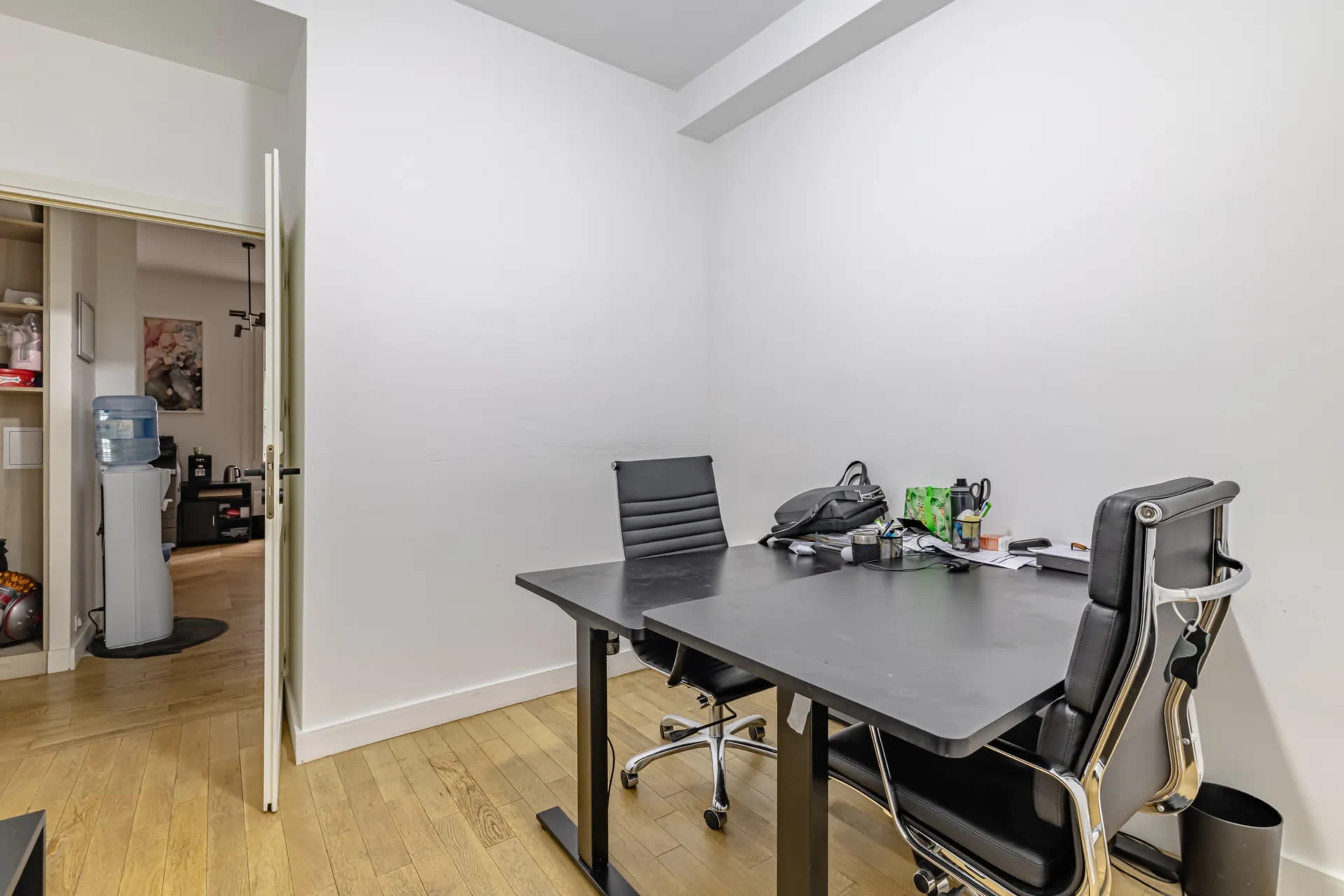 The image shows a minimalist office space featuring a black desk with two black chairs and a water cooler in the background.