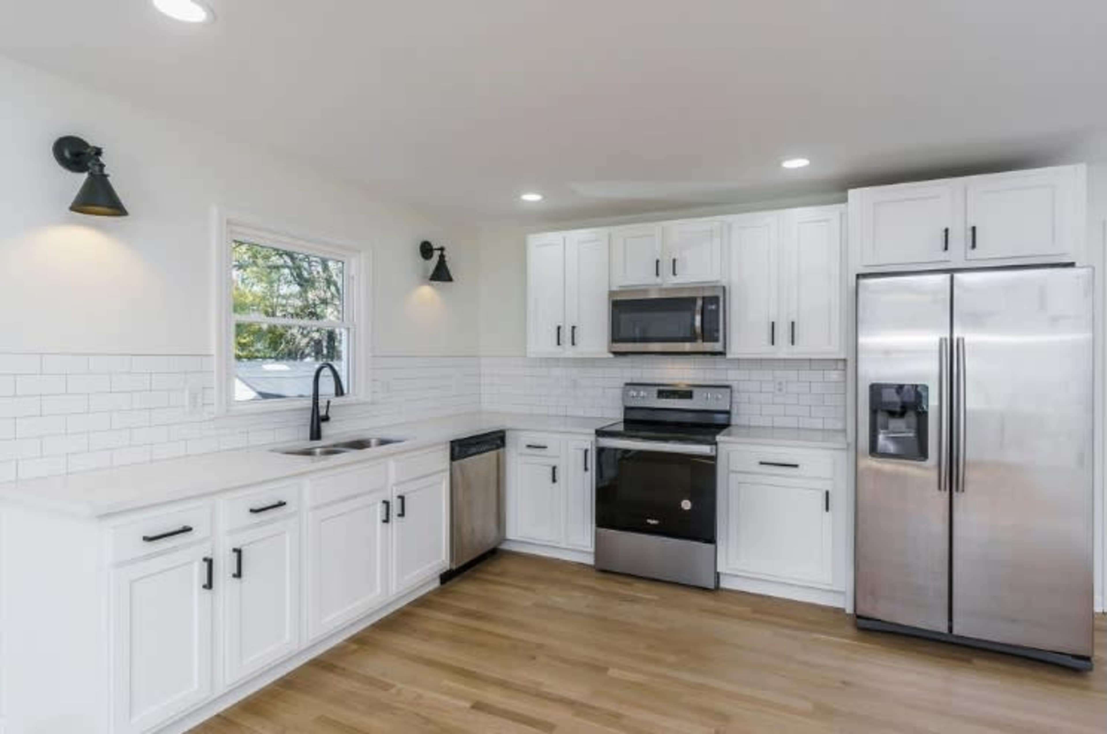 A modern kitchen features white cabinetry, stainless steel appliances, and a wood floor, with a large window providing natural light.