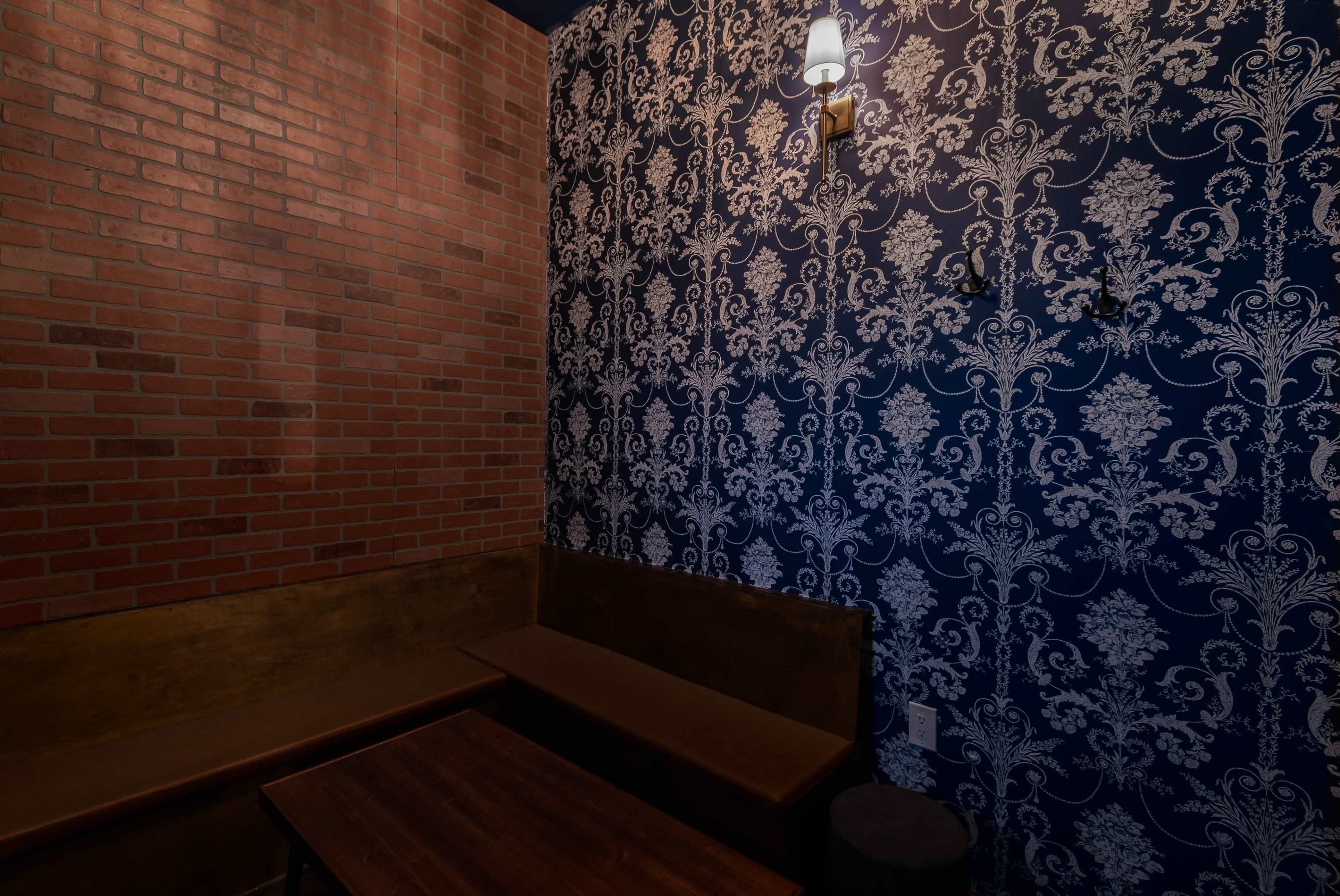 The image shows a corner of a room featuring a brick wall and a patterned blue wallpaper, with a wooden bench and a table.