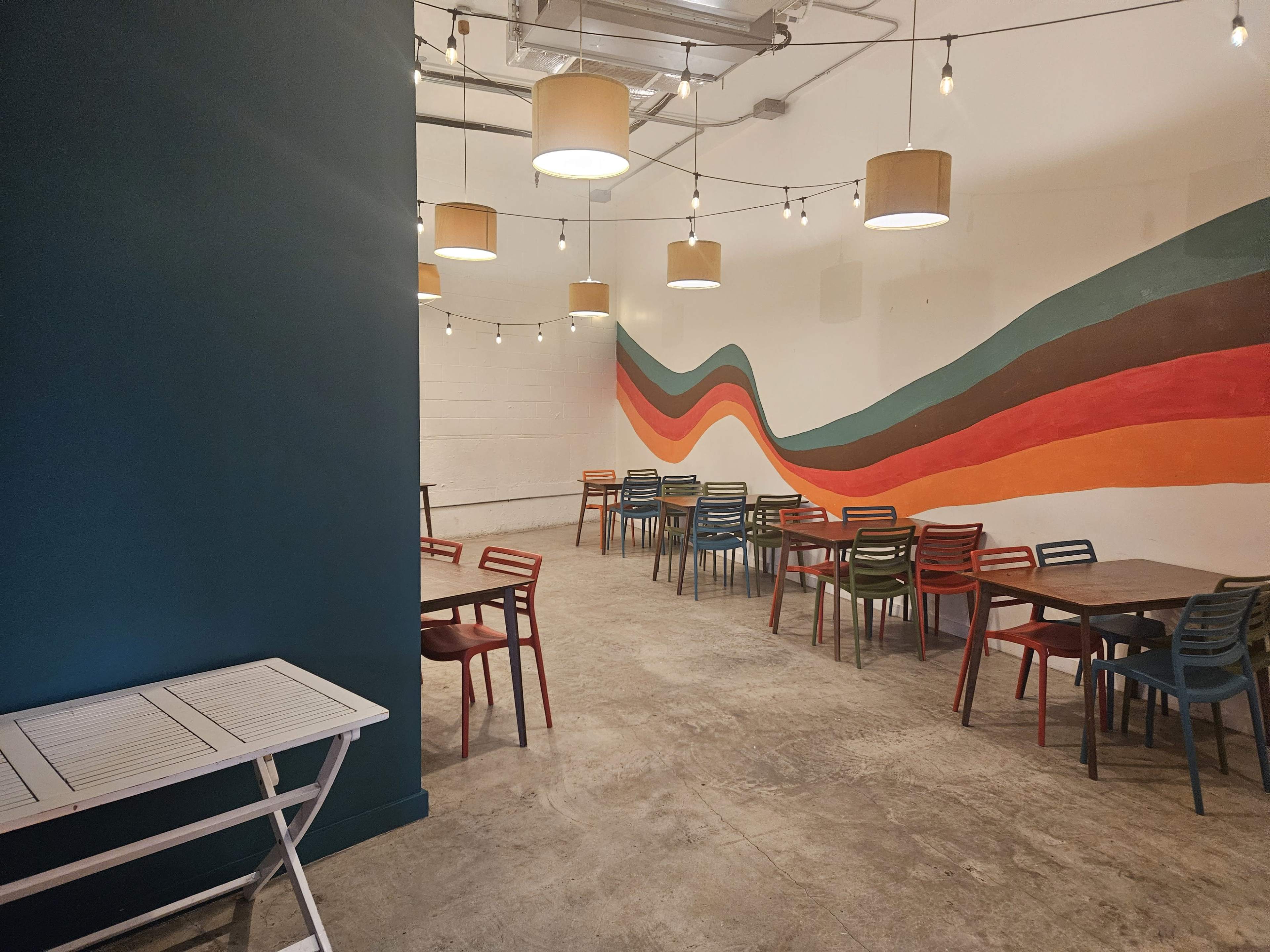 The image shows a spacious indoor dining area with colorful chairs and a wavy mural on the wall.