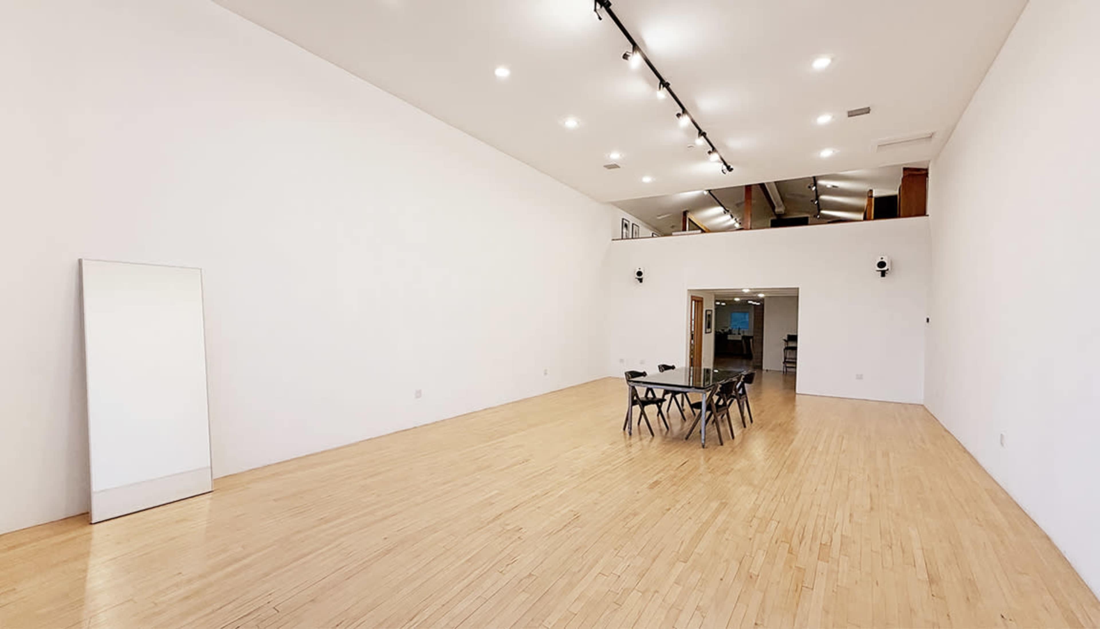 The image shows a spacious, empty room with wooden flooring, a large mirror on one wall, and a table set with chairs in the center.