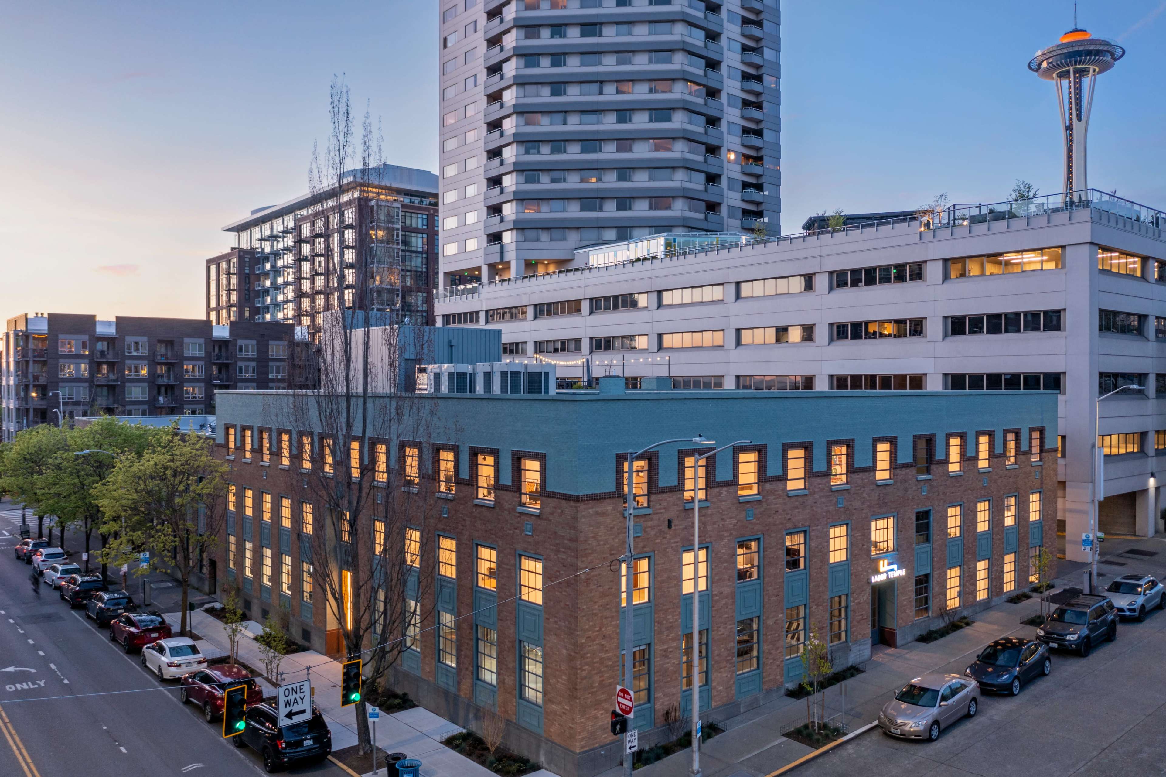 The image shows a multi-story building with a modern high-rise in the background, located in a city street lined with parked cars.