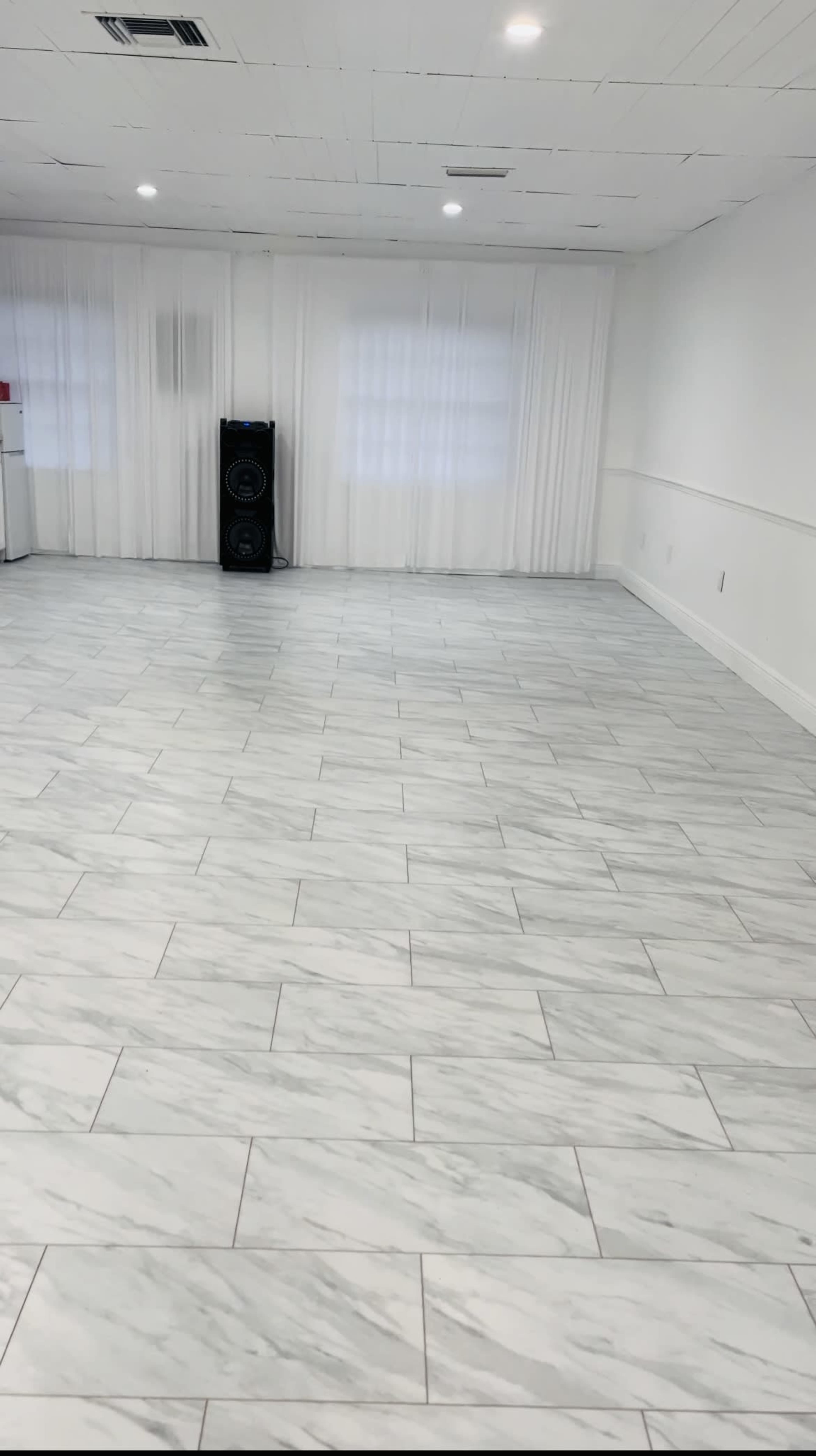 A spacious interior room with white marble tile flooring and large windows covered by sheer curtains.