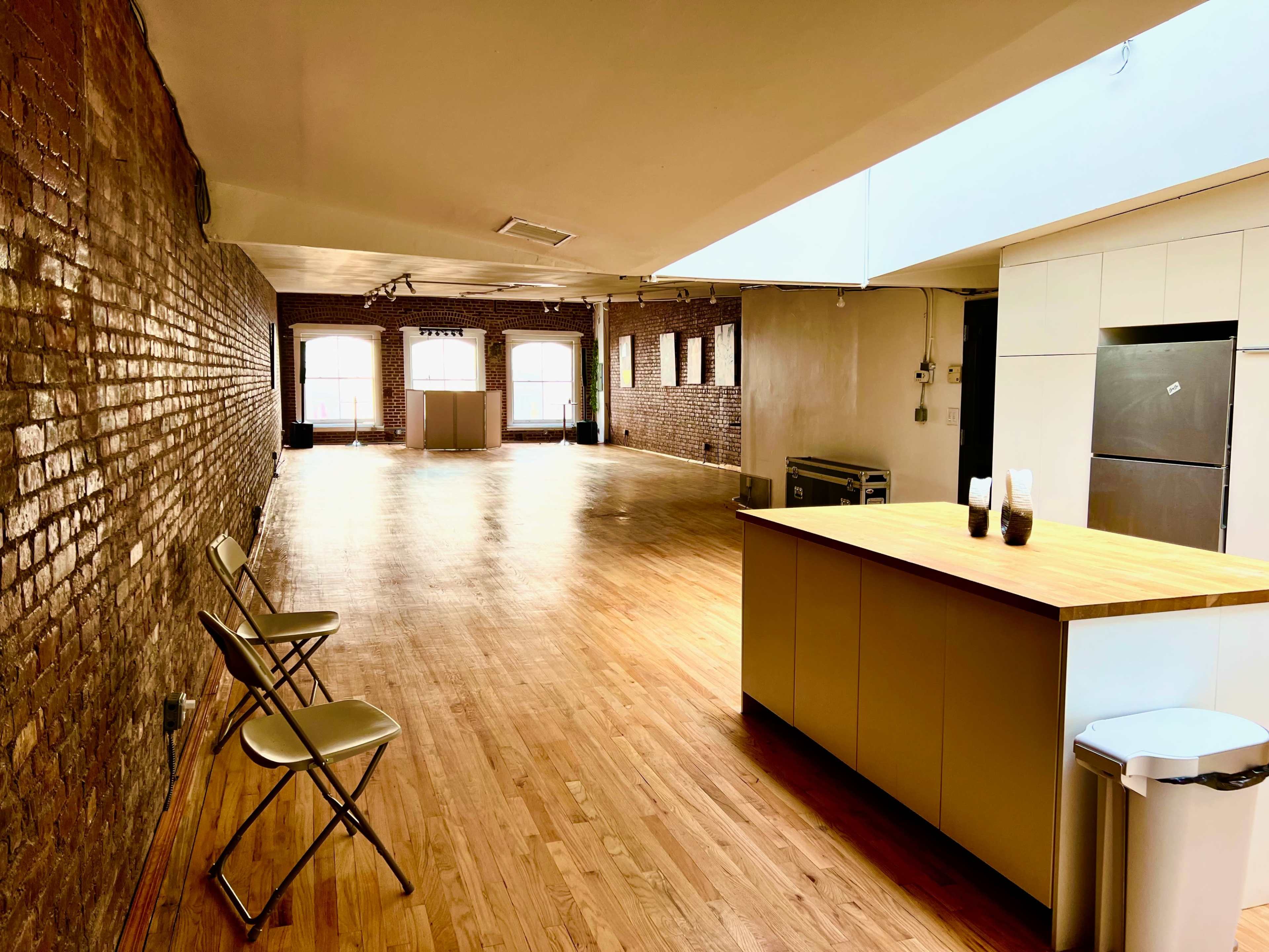 A spacious room with hardwood floors, exposed brick walls, and a kitchen area with a small island and metal chairs.
