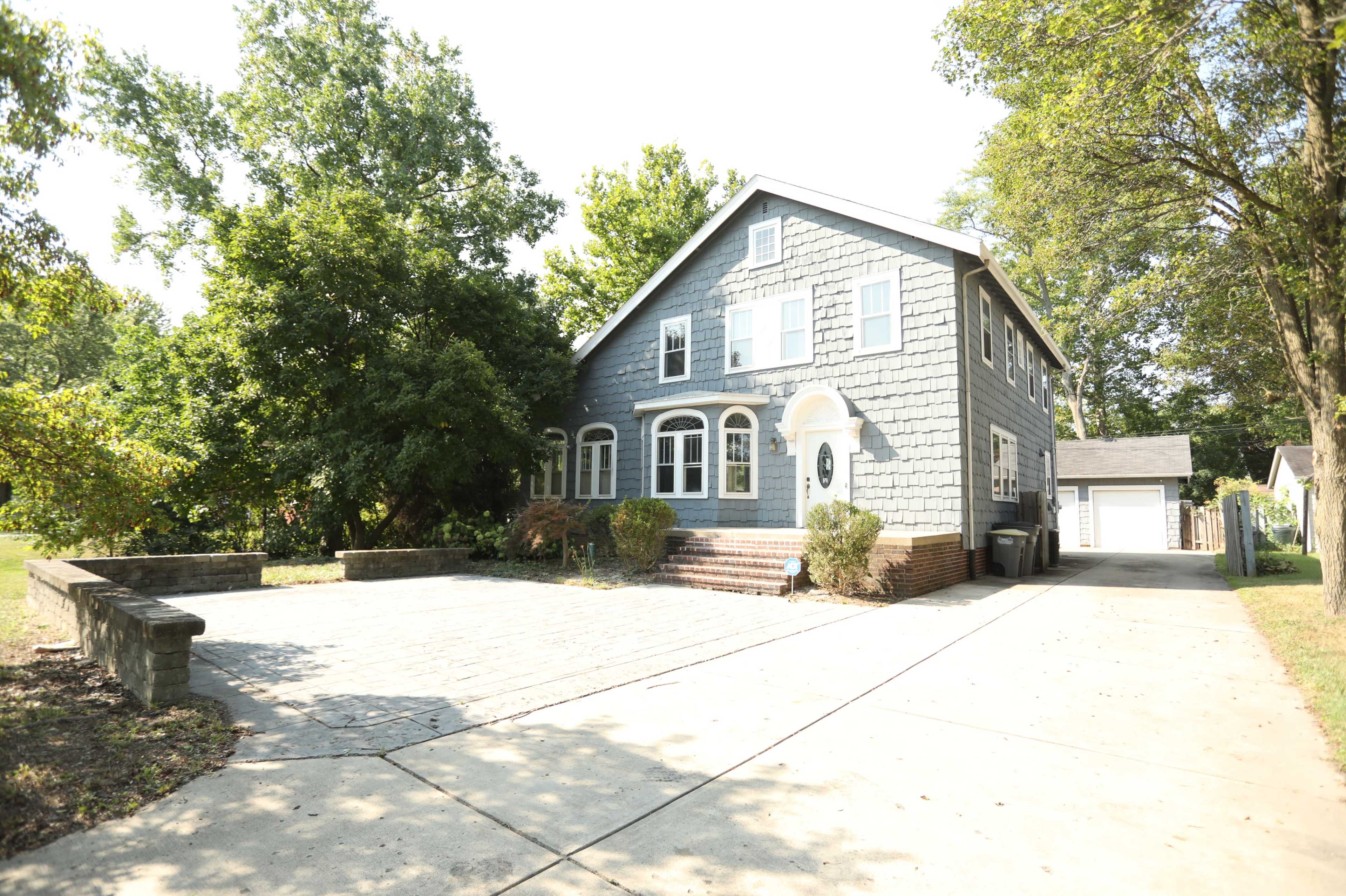 A large two-story house with gray siding and a spacious driveway is surrounded by trees and greenery.