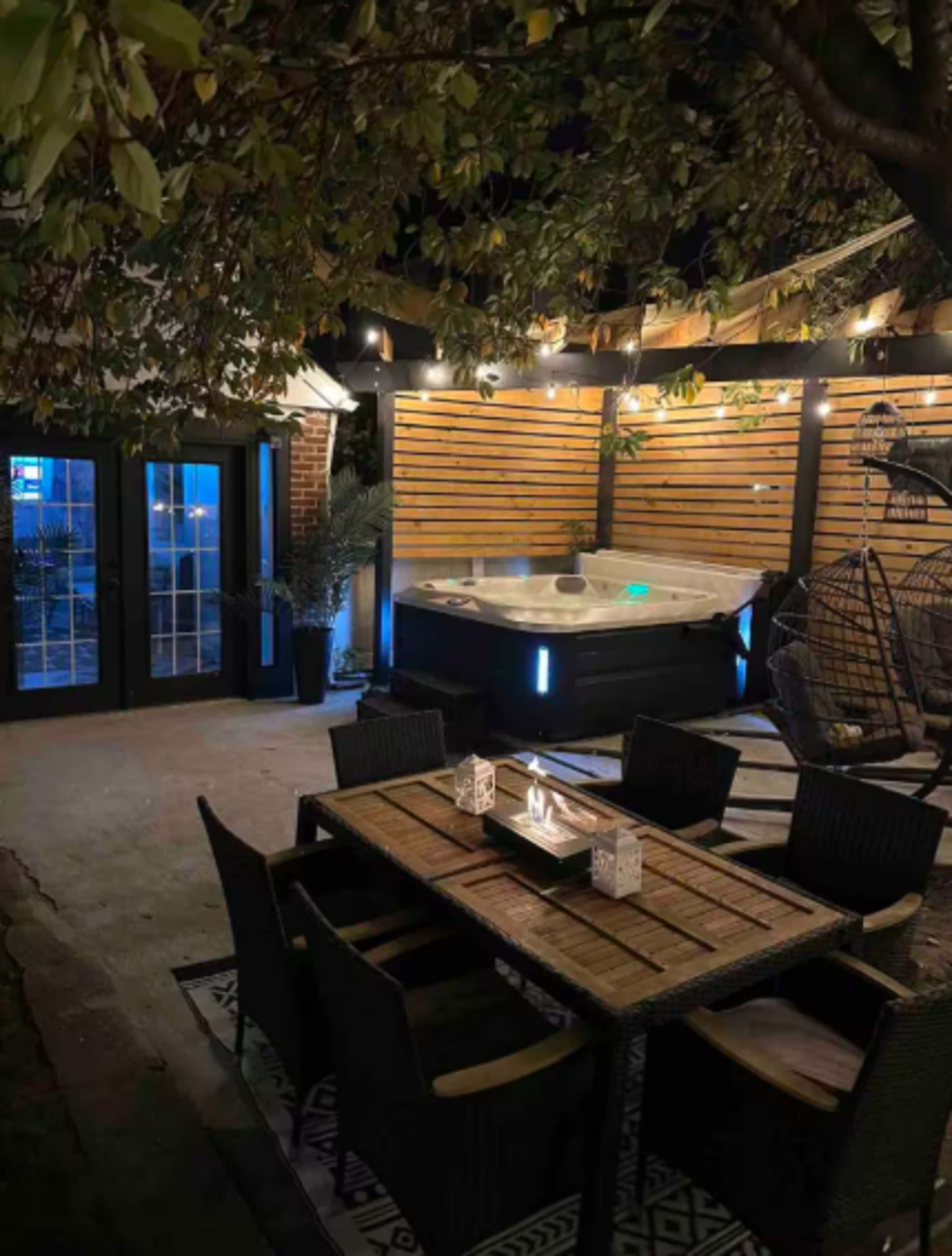 The image shows a dimly lit outdoor area featuring a hot tub, a dining table with chairs, and a backdrop of wooden privacy slats.