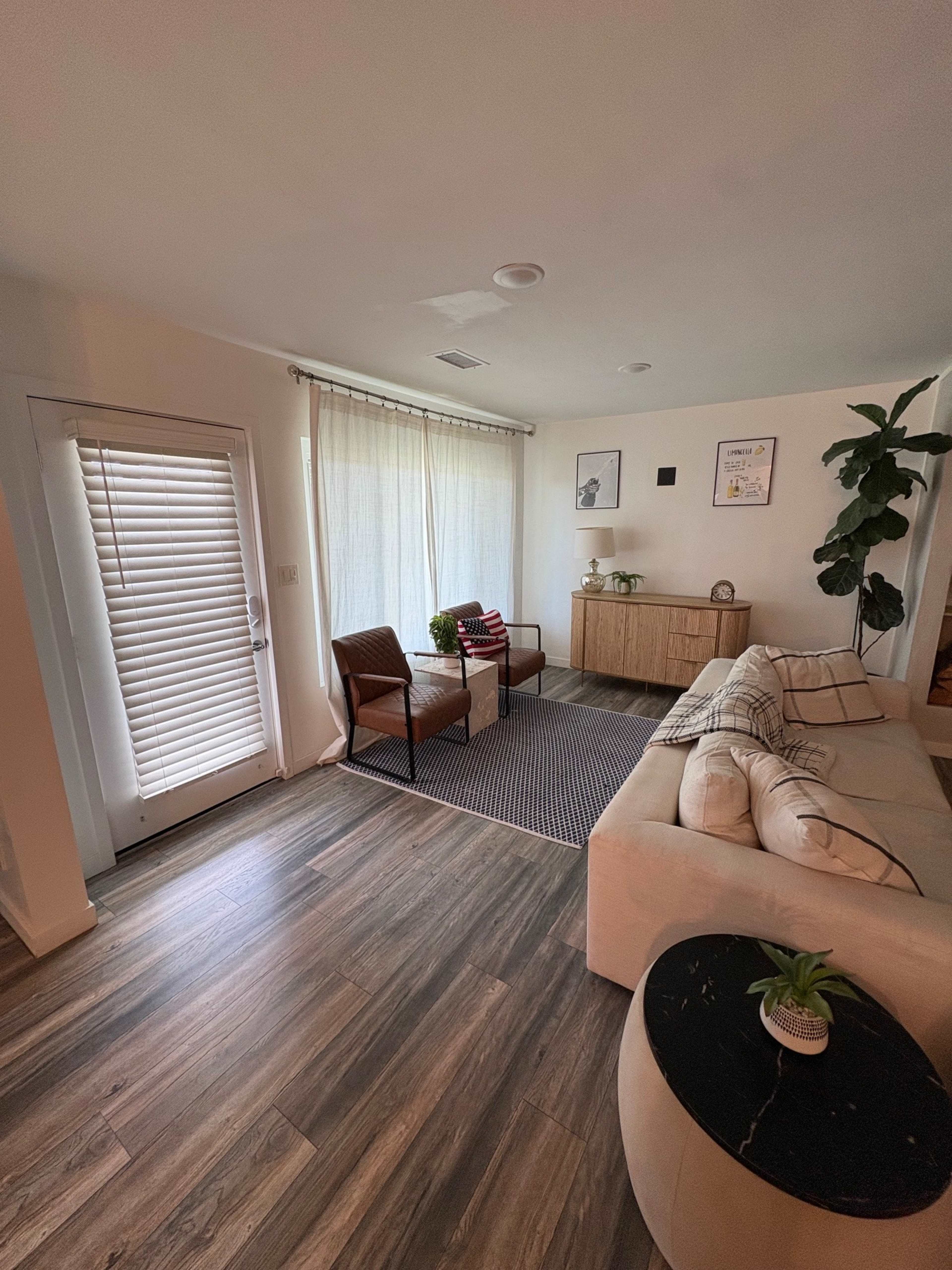 The image shows a modern living room with a beige sofa, two armchairs, a wooden sideboard, and a door leading outside, illuminated by natural light through sheer curtains.