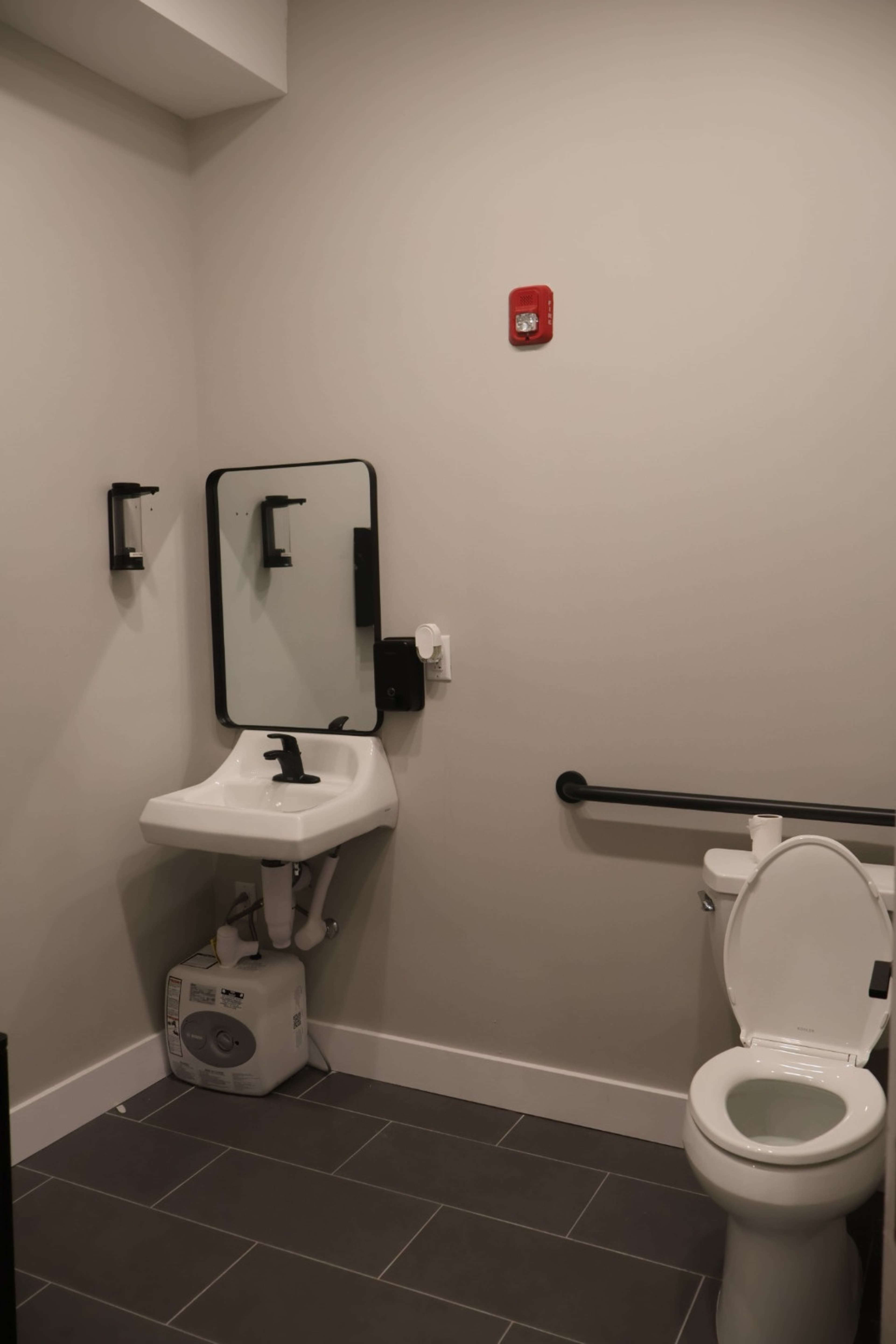 The image shows a small restroom featuring a toilet, a wall-mounted sink with a mirror, and a grab bar for accessibility.