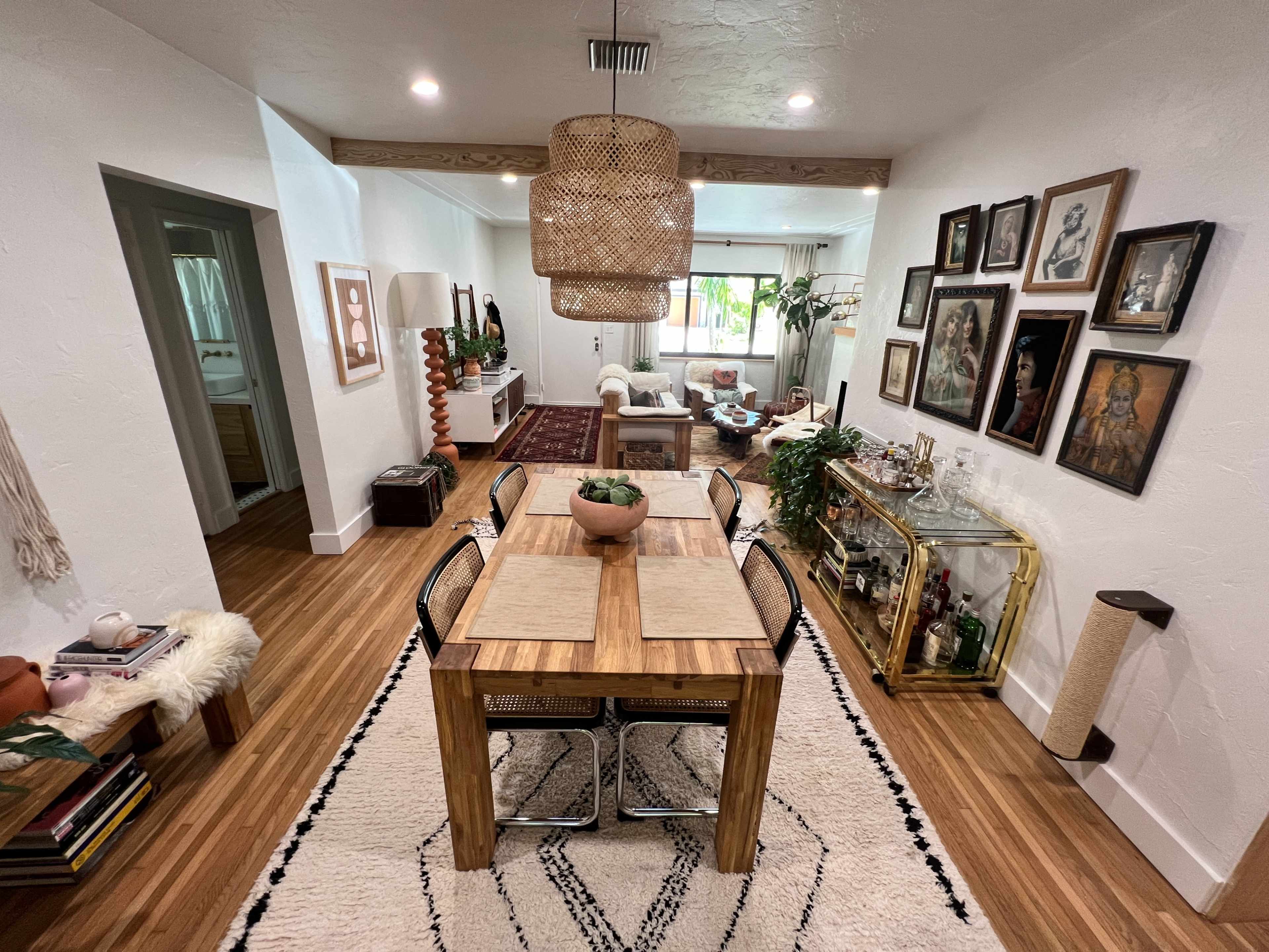 A modern dining room features a wooden table set for six, surrounded by woven chairs, with a decorative light fixture overhead and various artworks on the walls.