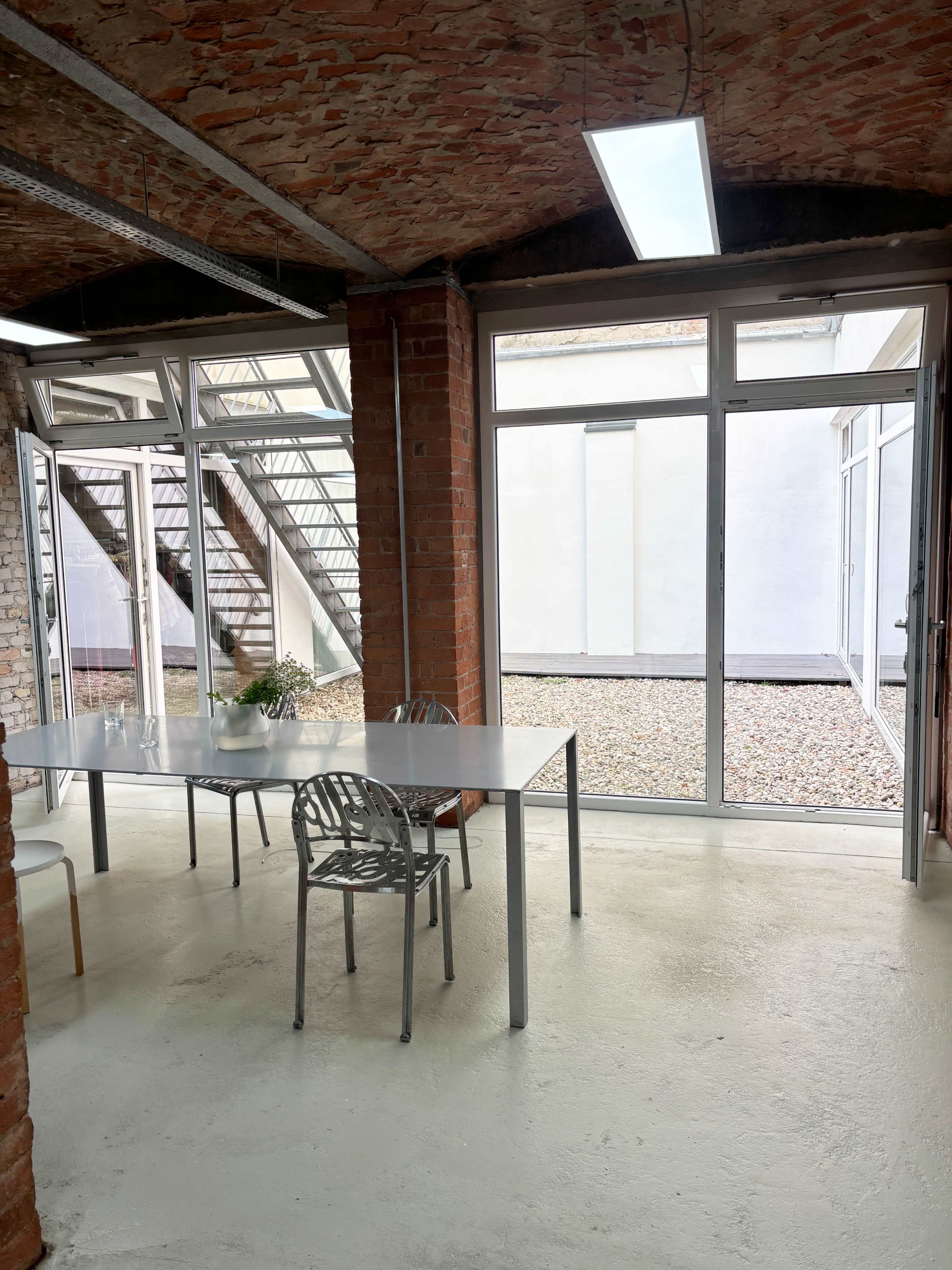 The image shows a minimalist office space with a long table and chairs, large windows, and a courtyard visible through the glass doors.