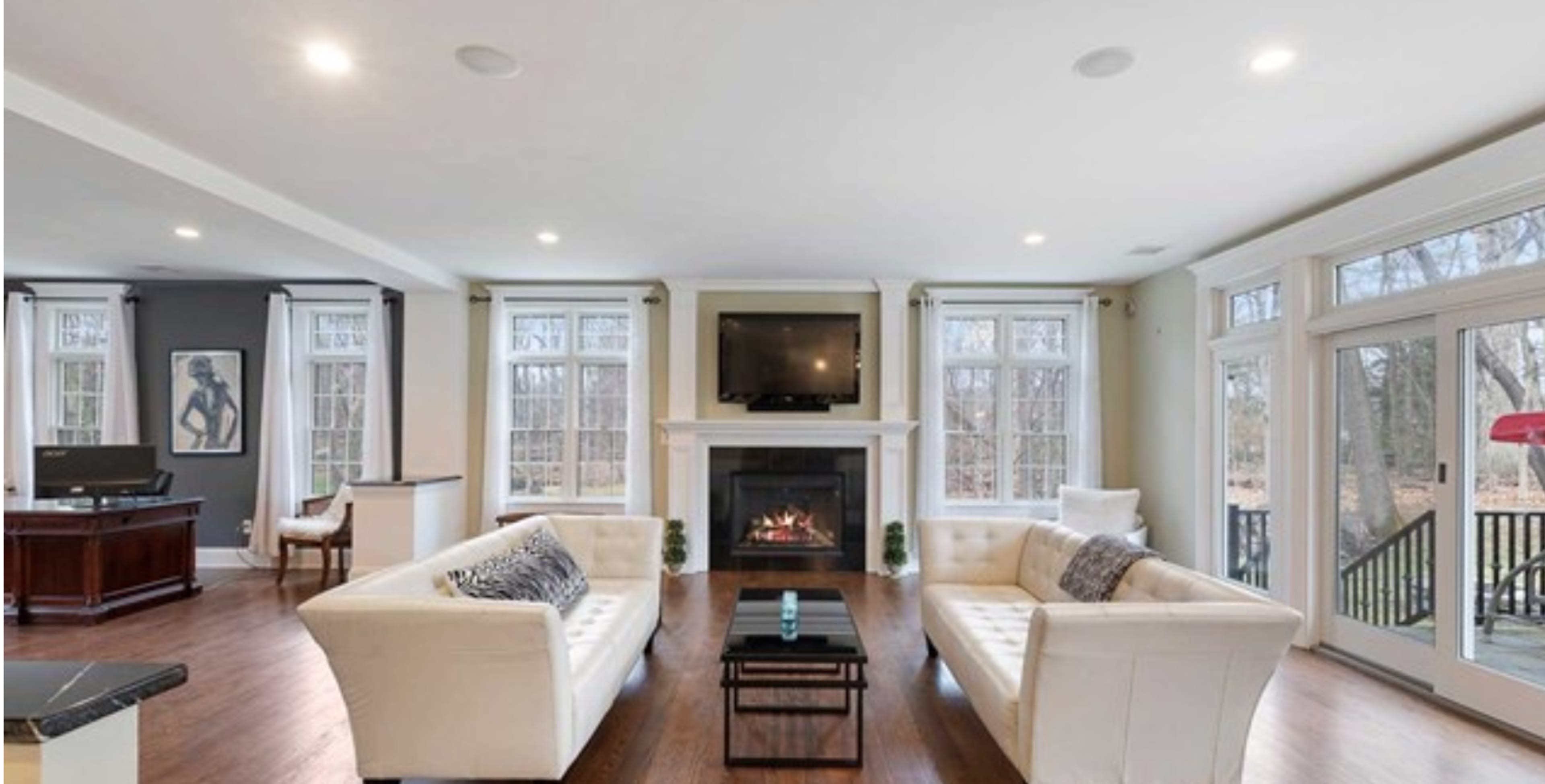 A modern living room features two white sofas facing a fireplace, with a television mounted above it and large windows providing a view of the outdoors.