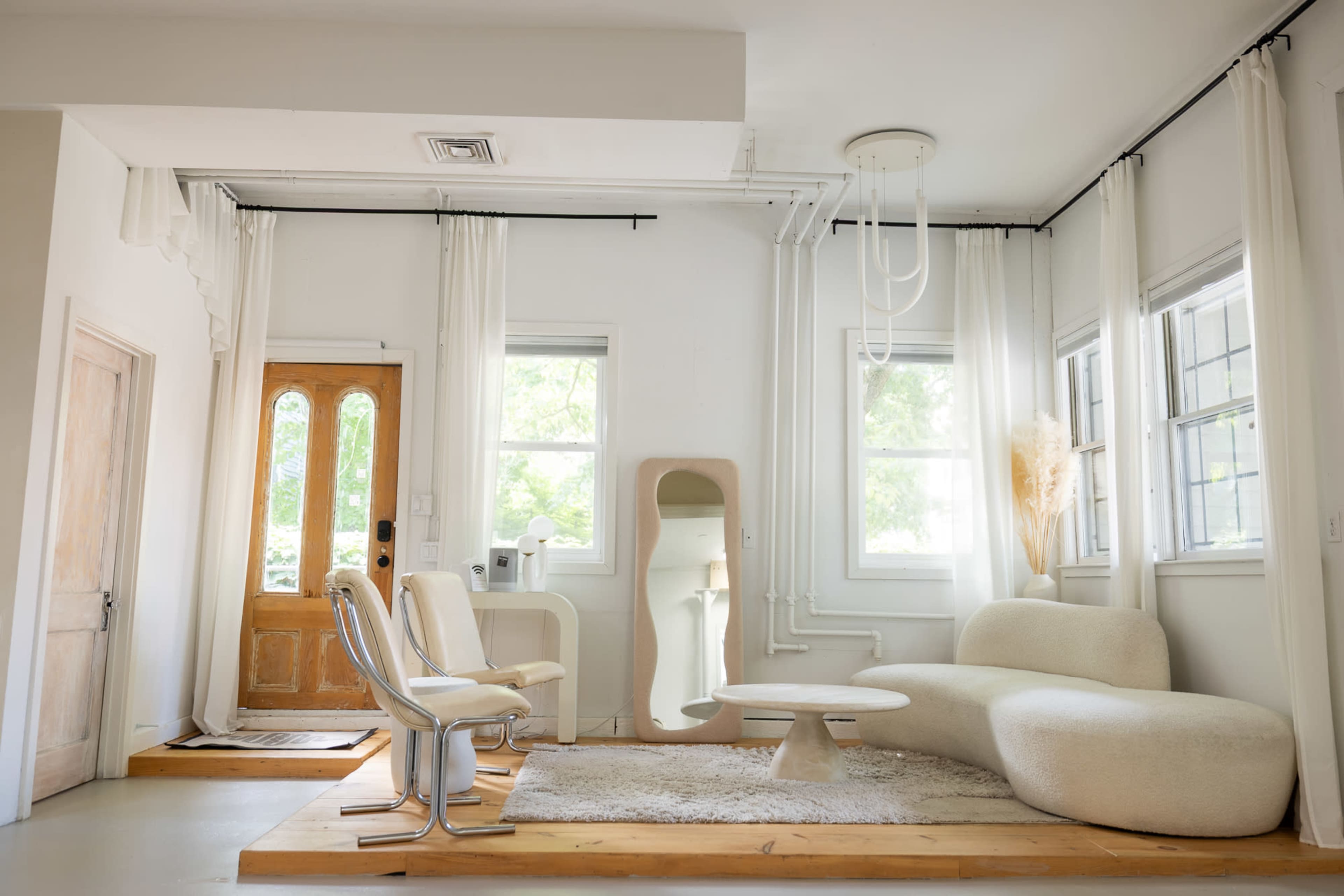 The image shows a bright, minimalist living room with a wooden floor, a curved sofa, a round table, and large windows draped with sheer curtains.