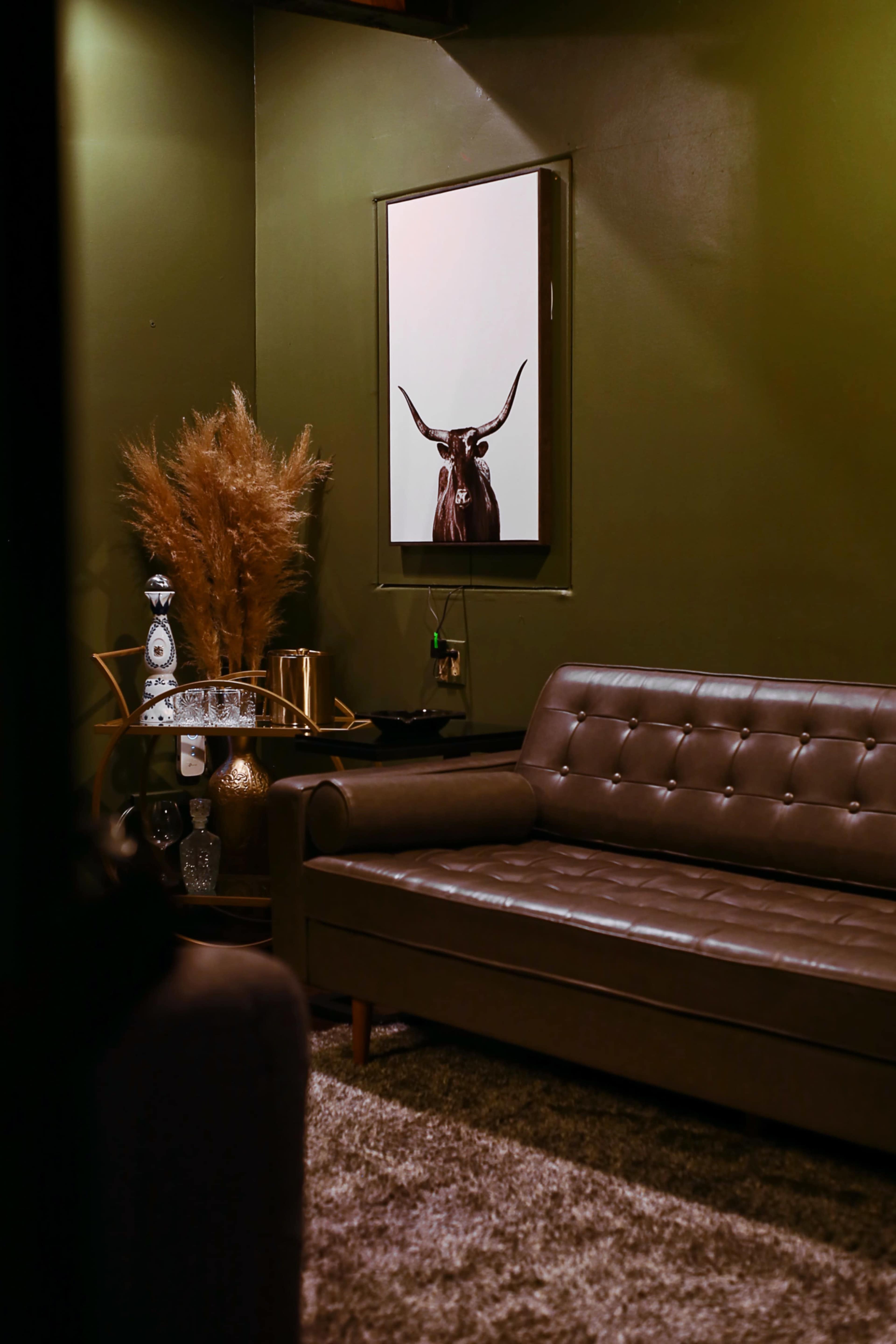 A dimly lit room with a brown leather sofa, a decorative mirror featuring a bull's head, and a wall adorned in dark green paint.
