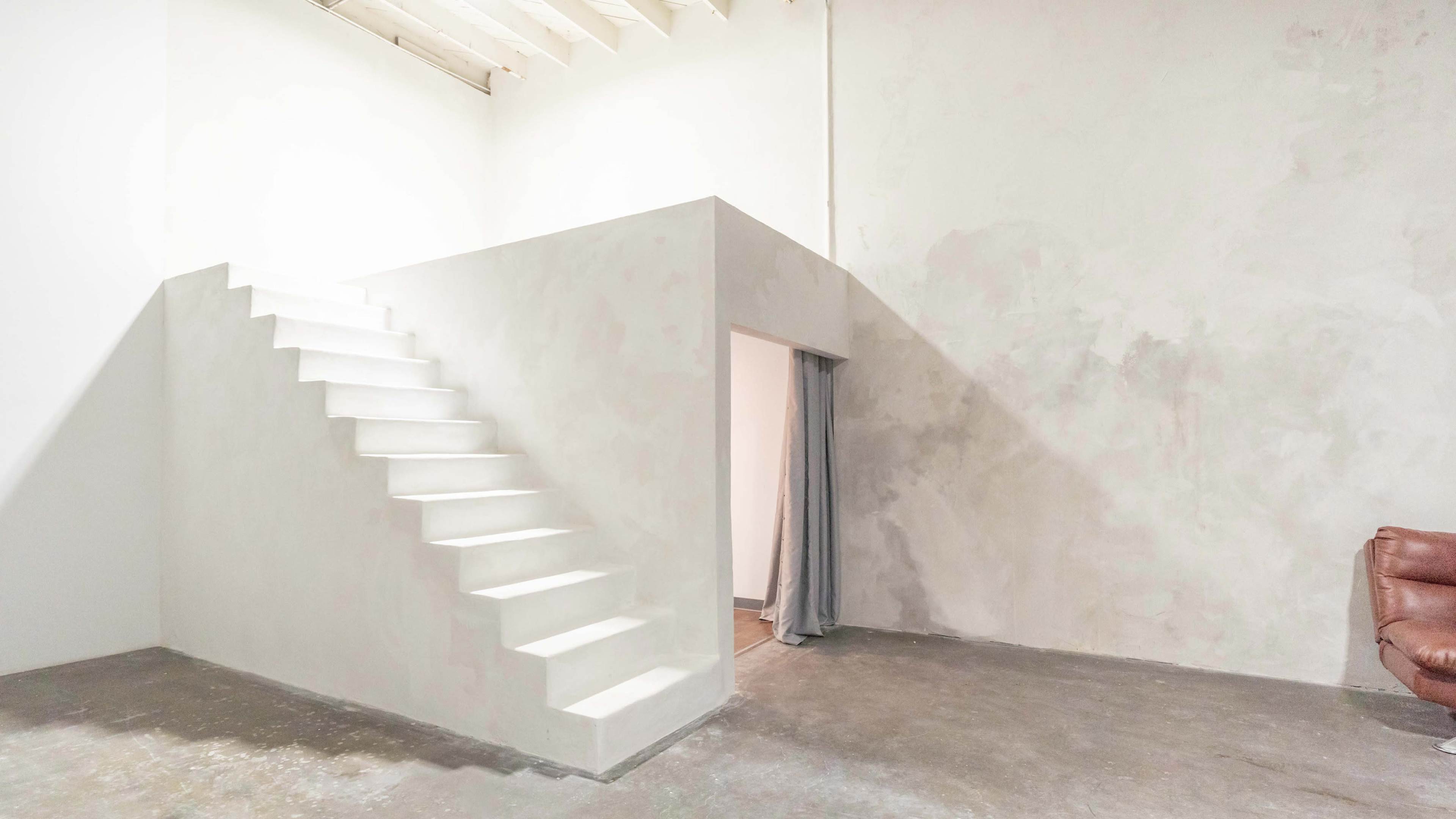 The image shows a minimalistic interior space featuring a white staircase leading to an elevated area, adjacent to a doorway with a gray curtain and a brown chair.