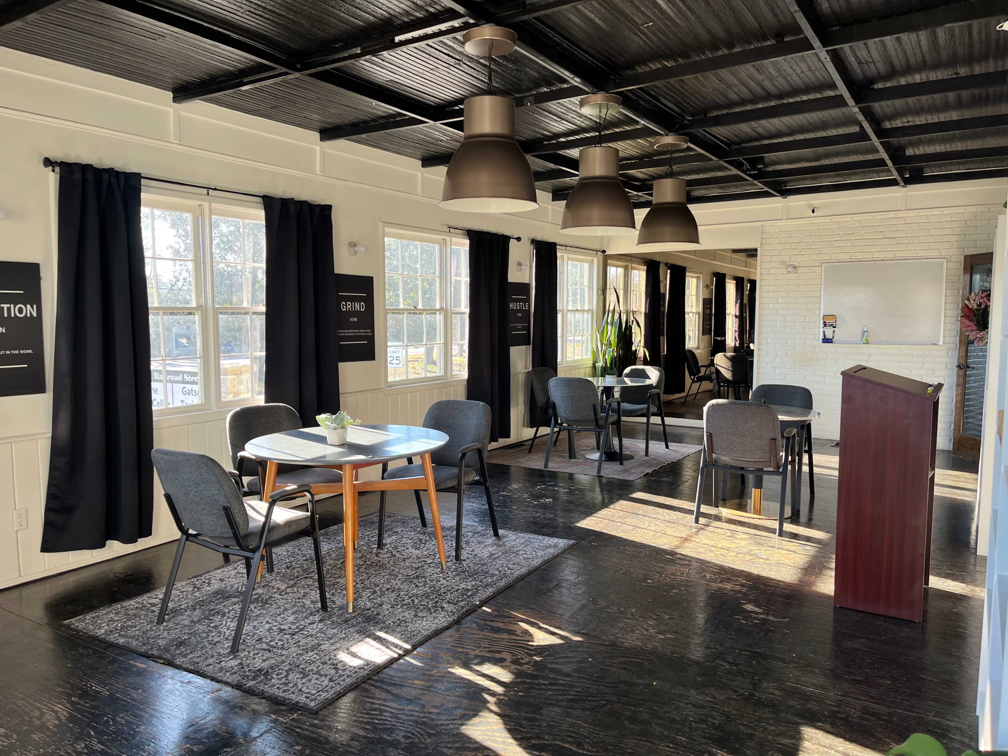 A sunlit room with several tables and chairs arranged around a central area, featuring large windows and a podium at one end.
