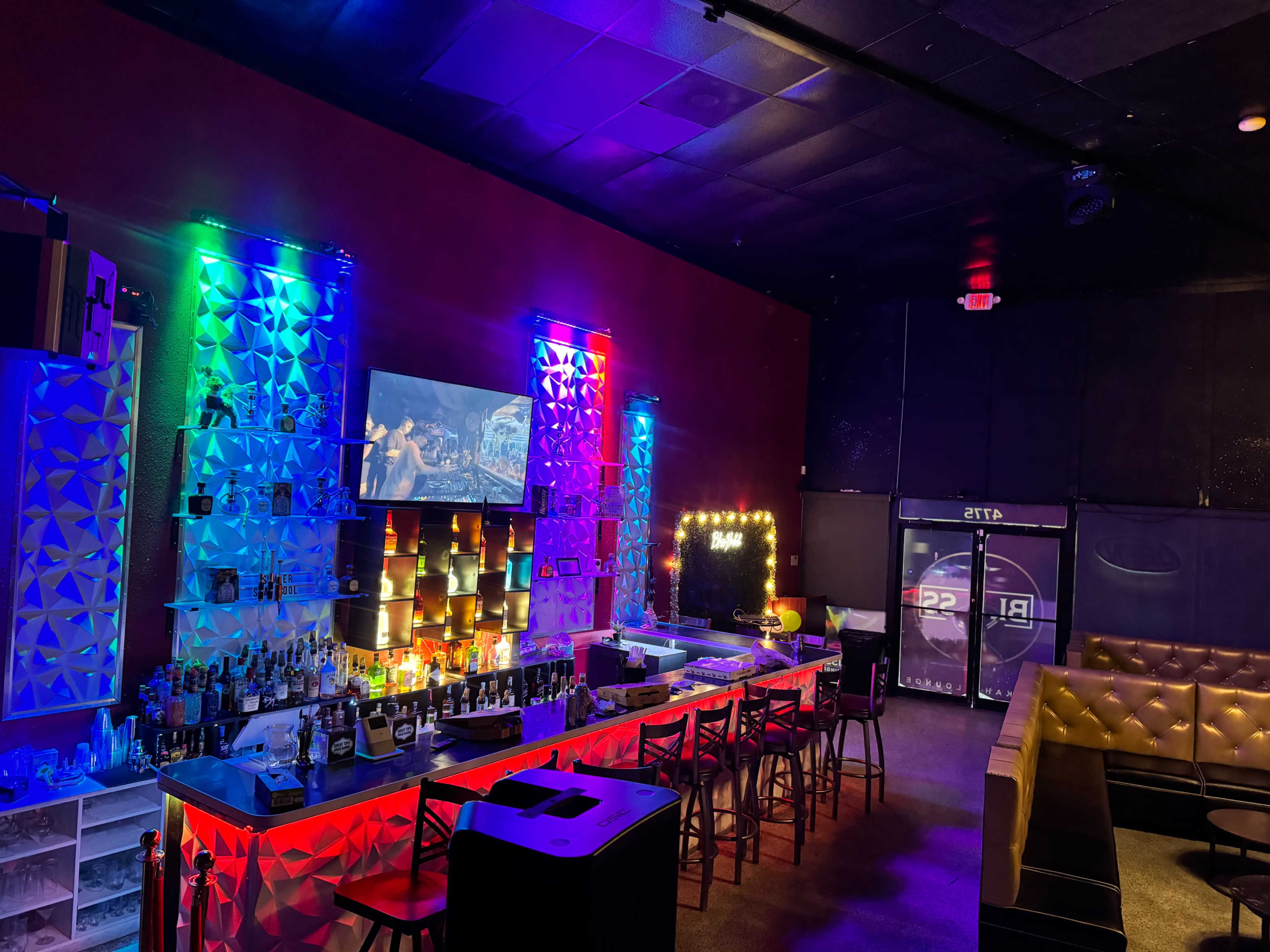 A bar area with illuminated shelves displaying various bottles, a large screen on the wall, and a seating area with a couch.