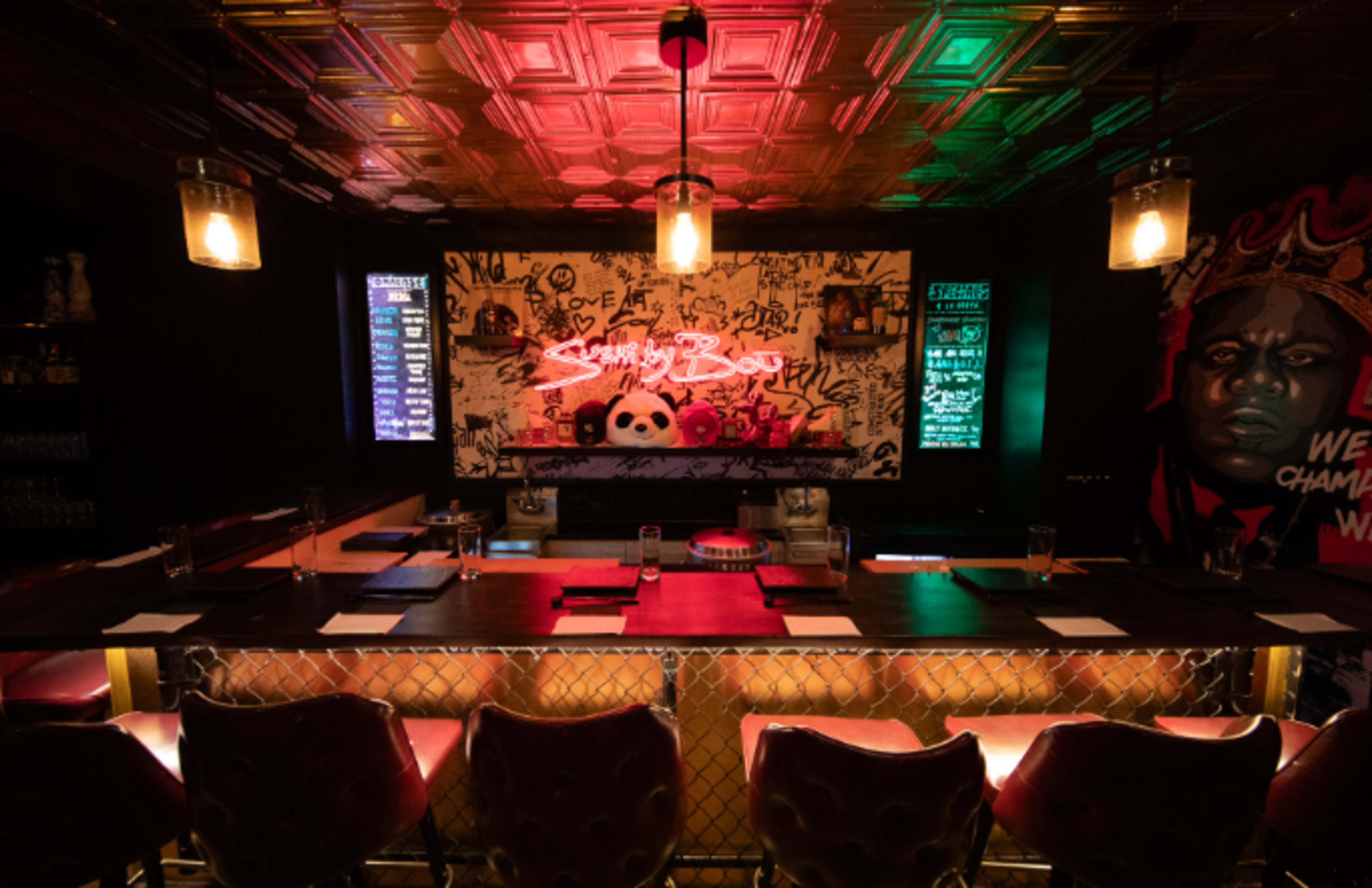 The image shows a dimly lit bar with neon graffiti art on the wall and seating along a long wooden counter.