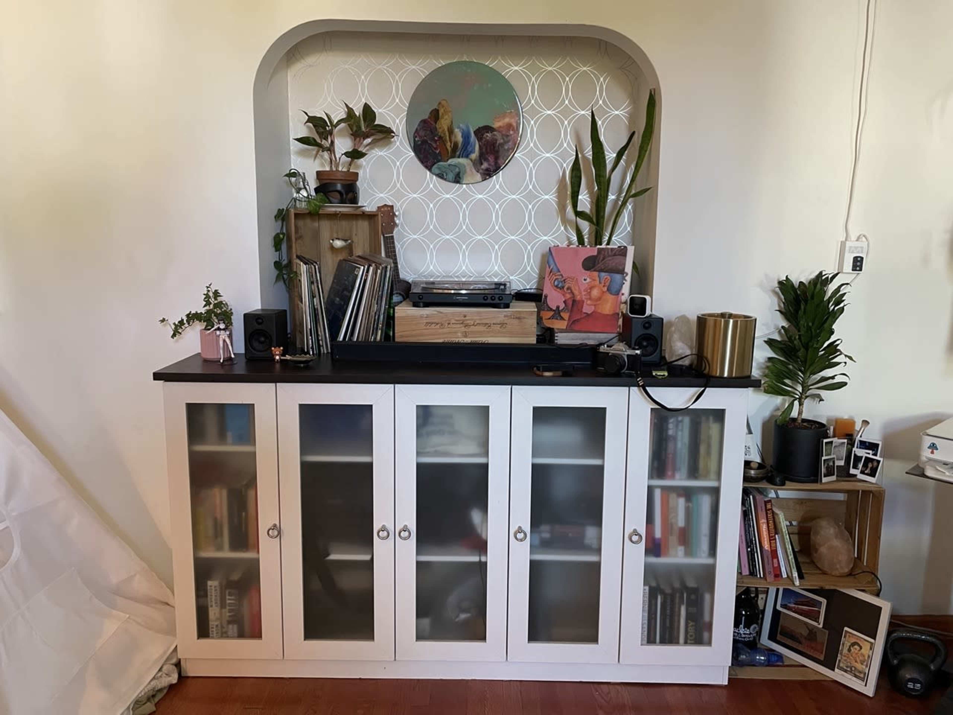 A white sideboard with glass-front cabinets displays books, plants, and record player equipment against a patterned backdrop.