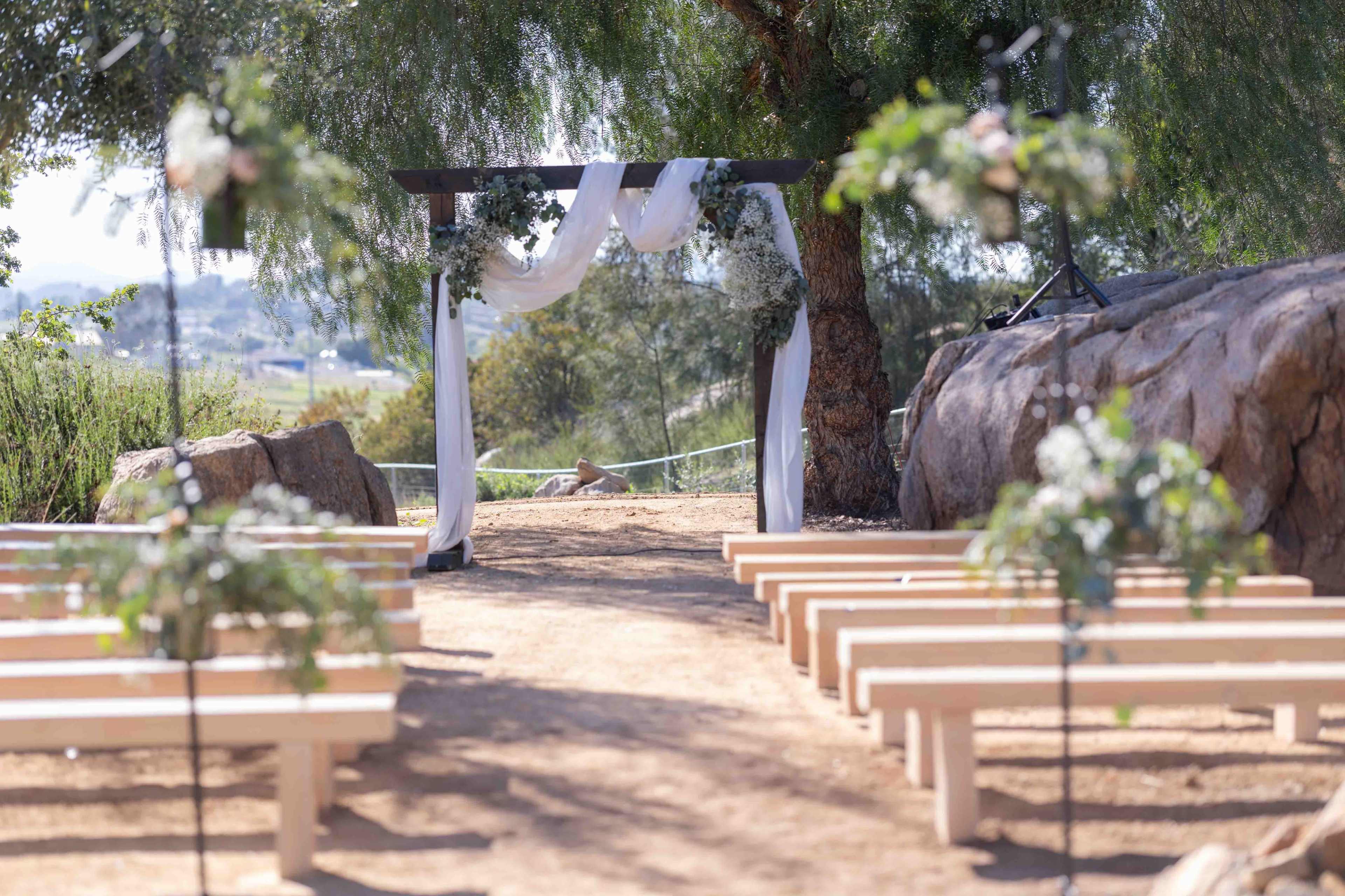 The image shows an outdoor wedding setup with white benches arranged in rows facing a decorated archway draped with fabric, surrounded by greenery.