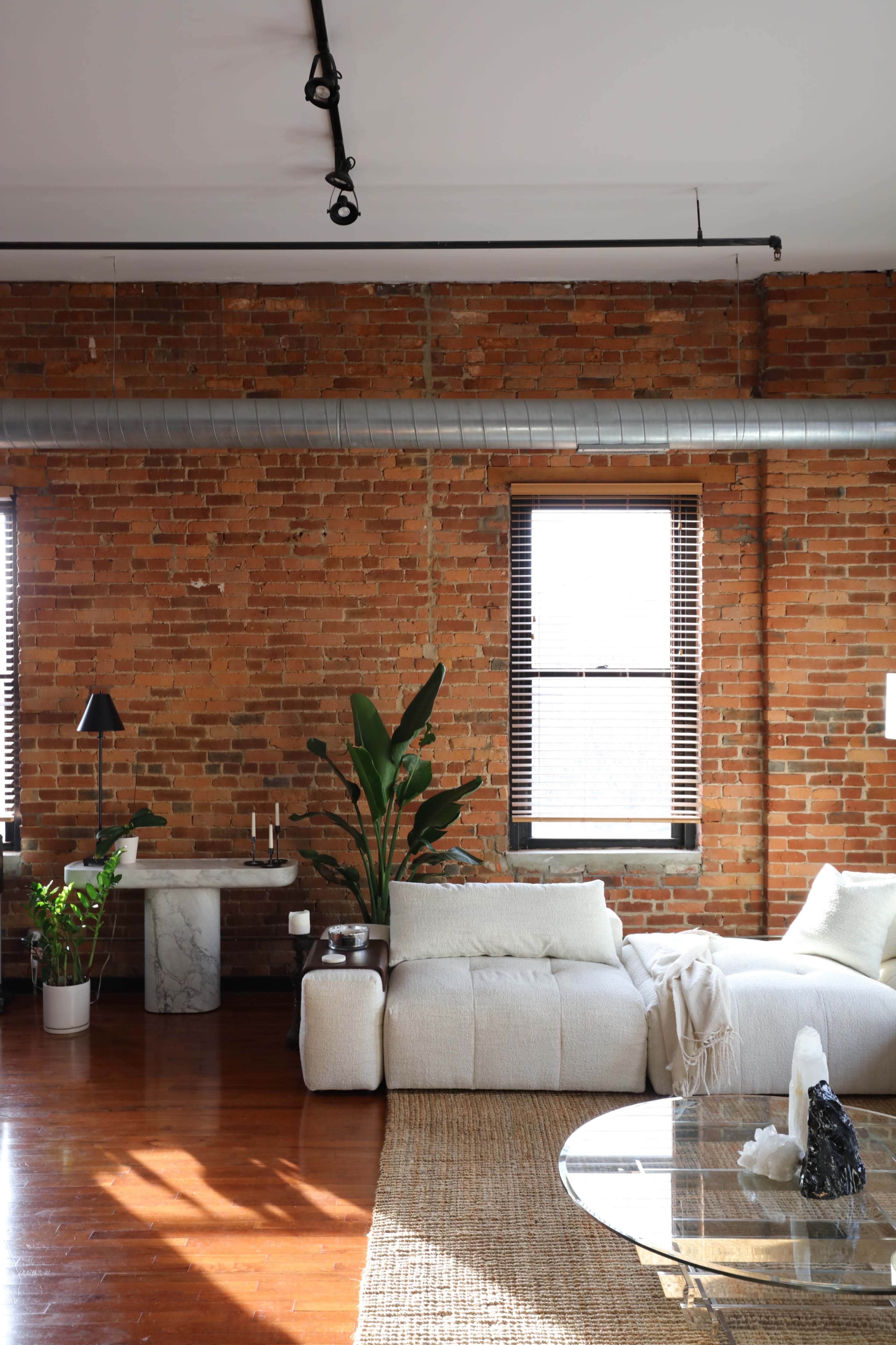 A modern living room with exposed brick walls, large windows, a light-colored sofa, a marble side table, and indoor plants.