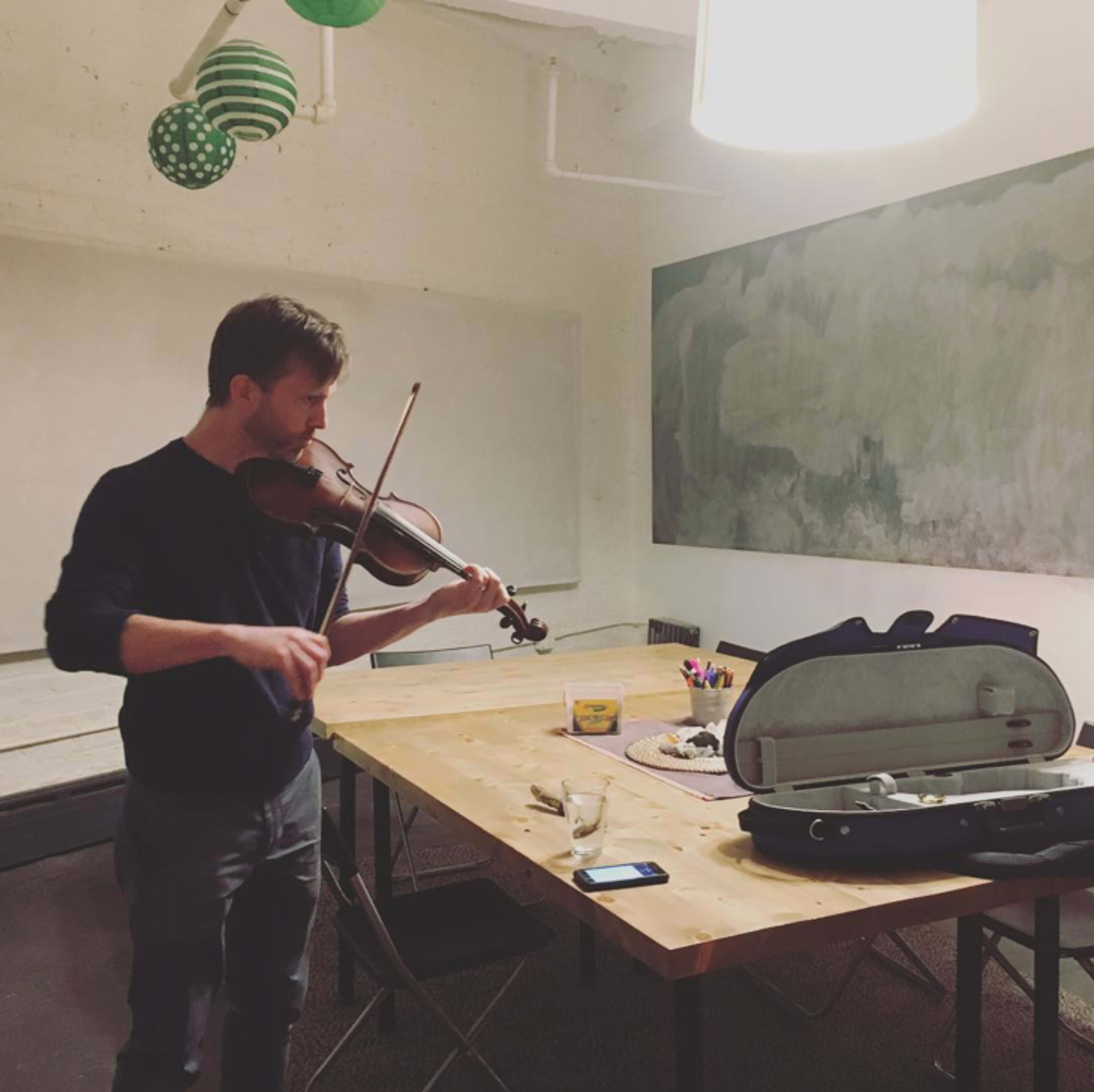 A man plays the violin in a room with a large table and a blank wall.