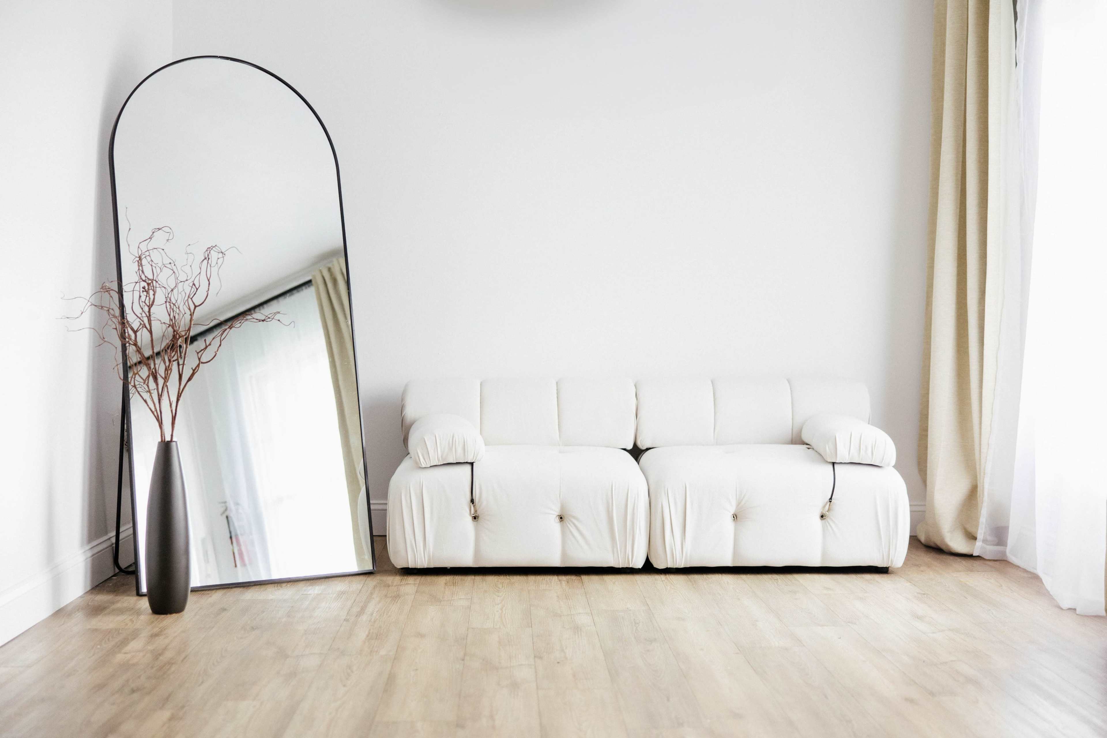 A modern living room features a white sofa beside a large, arched mirror and a tall vase with dried branches.