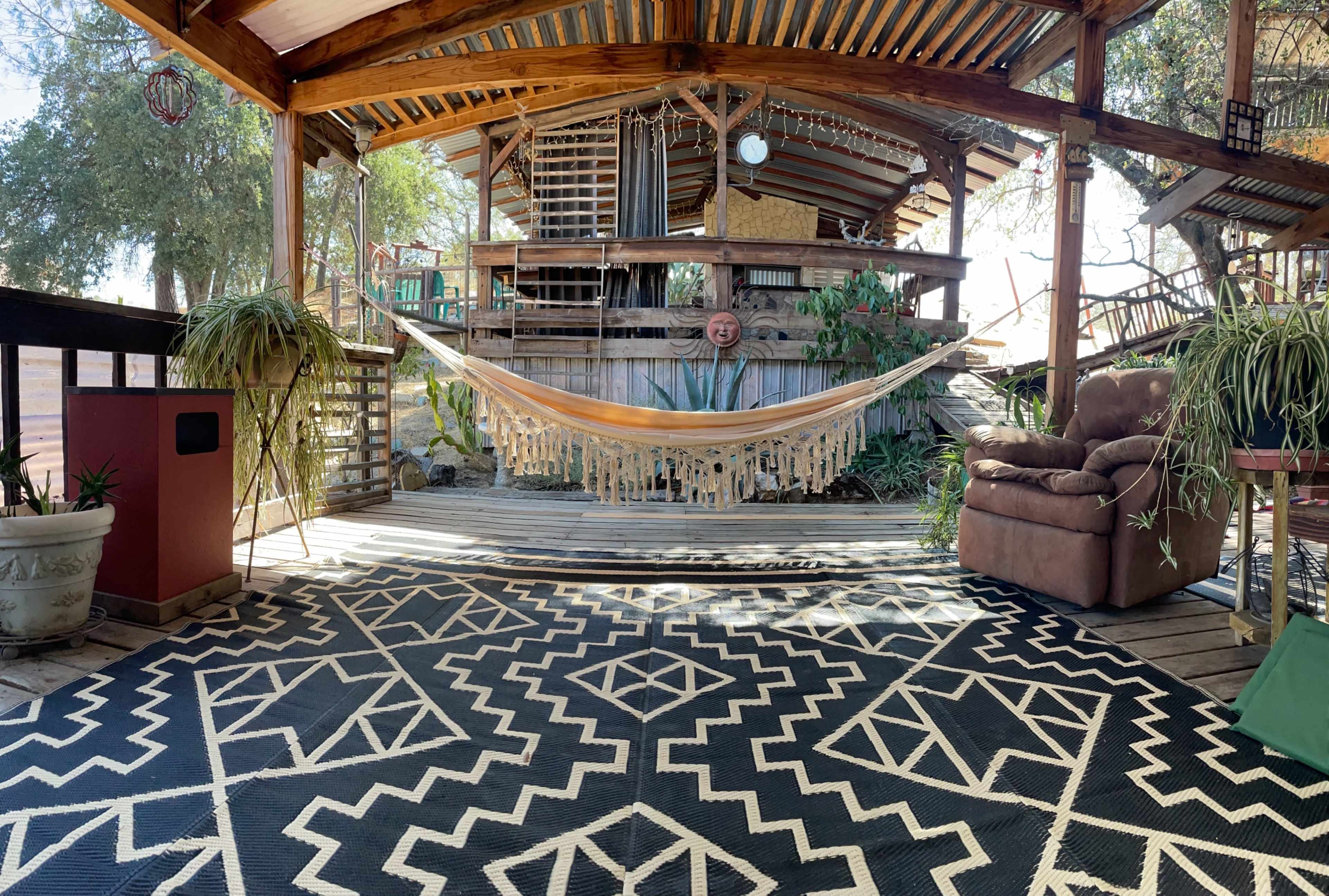 A wooden deck with a hammock, potted plants, and a patterned rug under a canopy of trees.